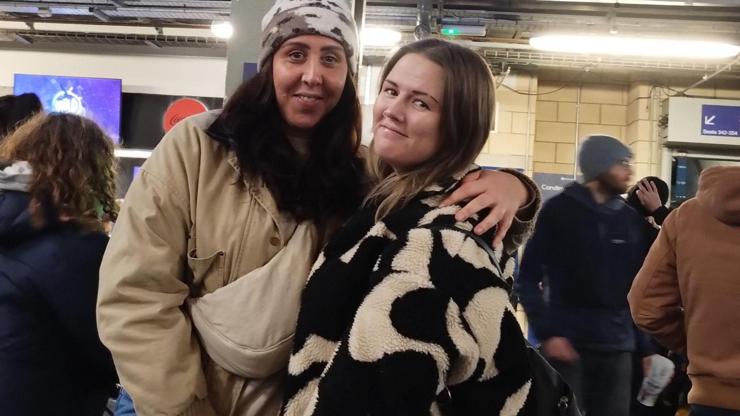 Grace Goddard (left) and Holly Way (right) are standing together with people around them. They are both wearing coats and Ms Goddard is wearing a hat. There are screens on the walls behind them.