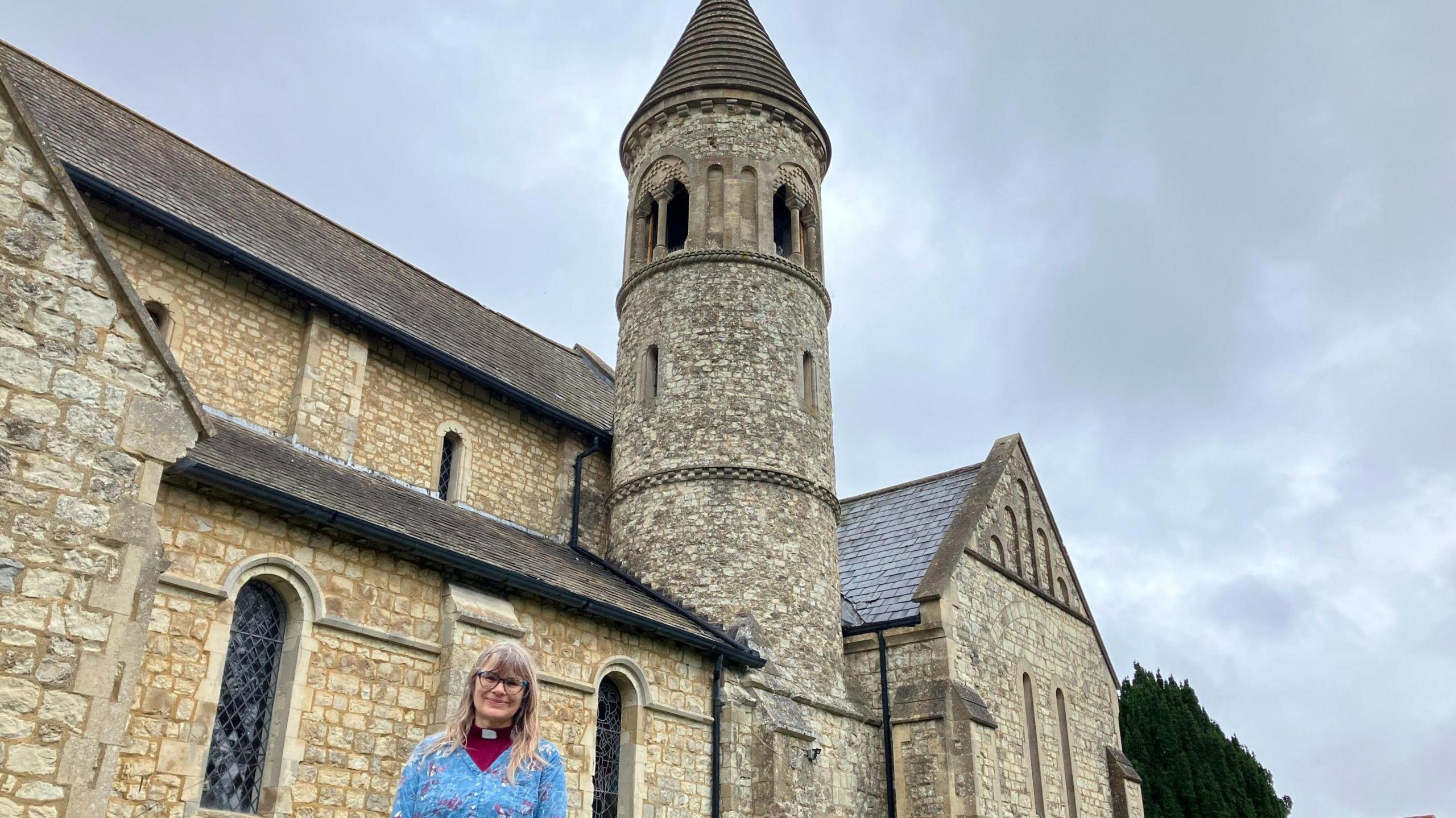 The exterior of St John's church in Hale, a grey building with a tower topped by a turret, built in the Romano architectural style, with the Rev Stella Wiseman, a middle aged woman with long fair hair, glasses, a clerical collar and a bright blue jumper, standing in front of it.