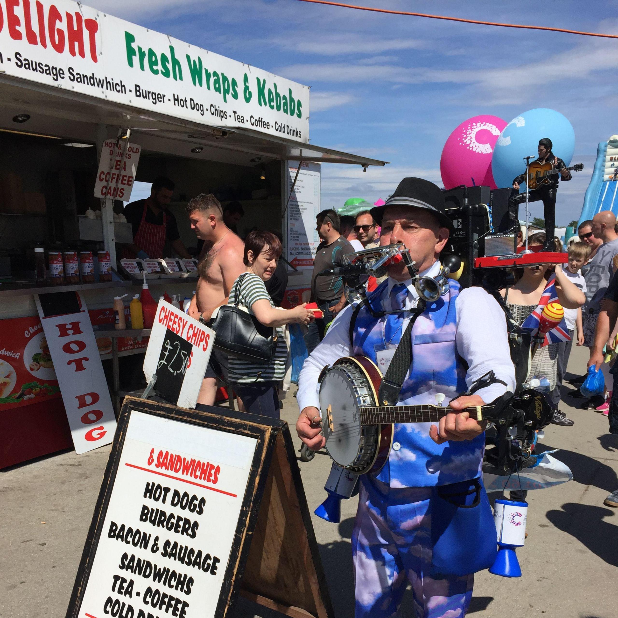 A man dressed in a blue and white suit with white clouds on it is wearing a black hat and playing a circular instrument with another instrument in his mouth. He has a cancer research pot next to him. You can also see a 'fresh wraps and kebabs' stall.