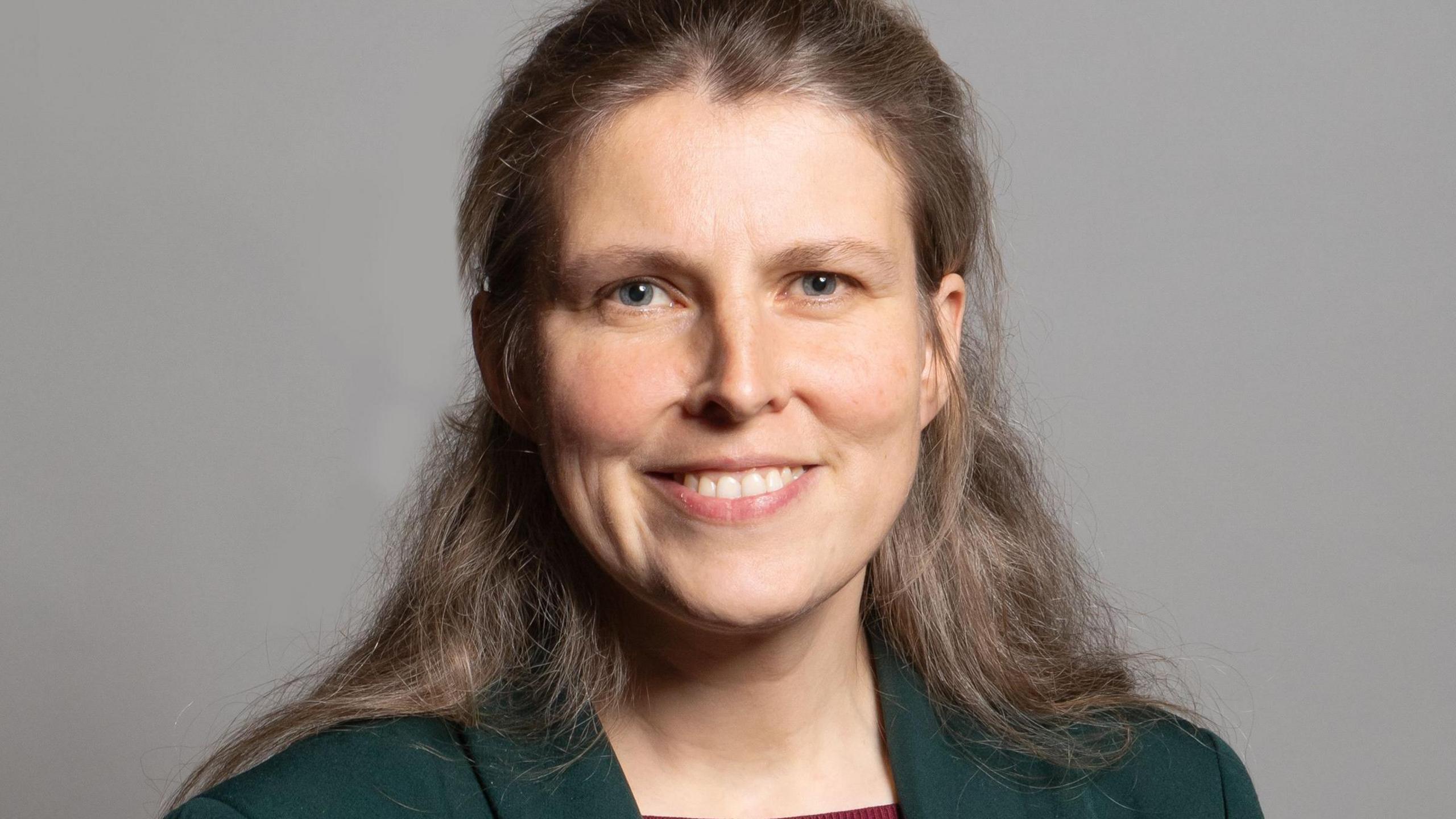 A woman with long light brown hair wearing a dark blazer. She stands in front of a grey background.