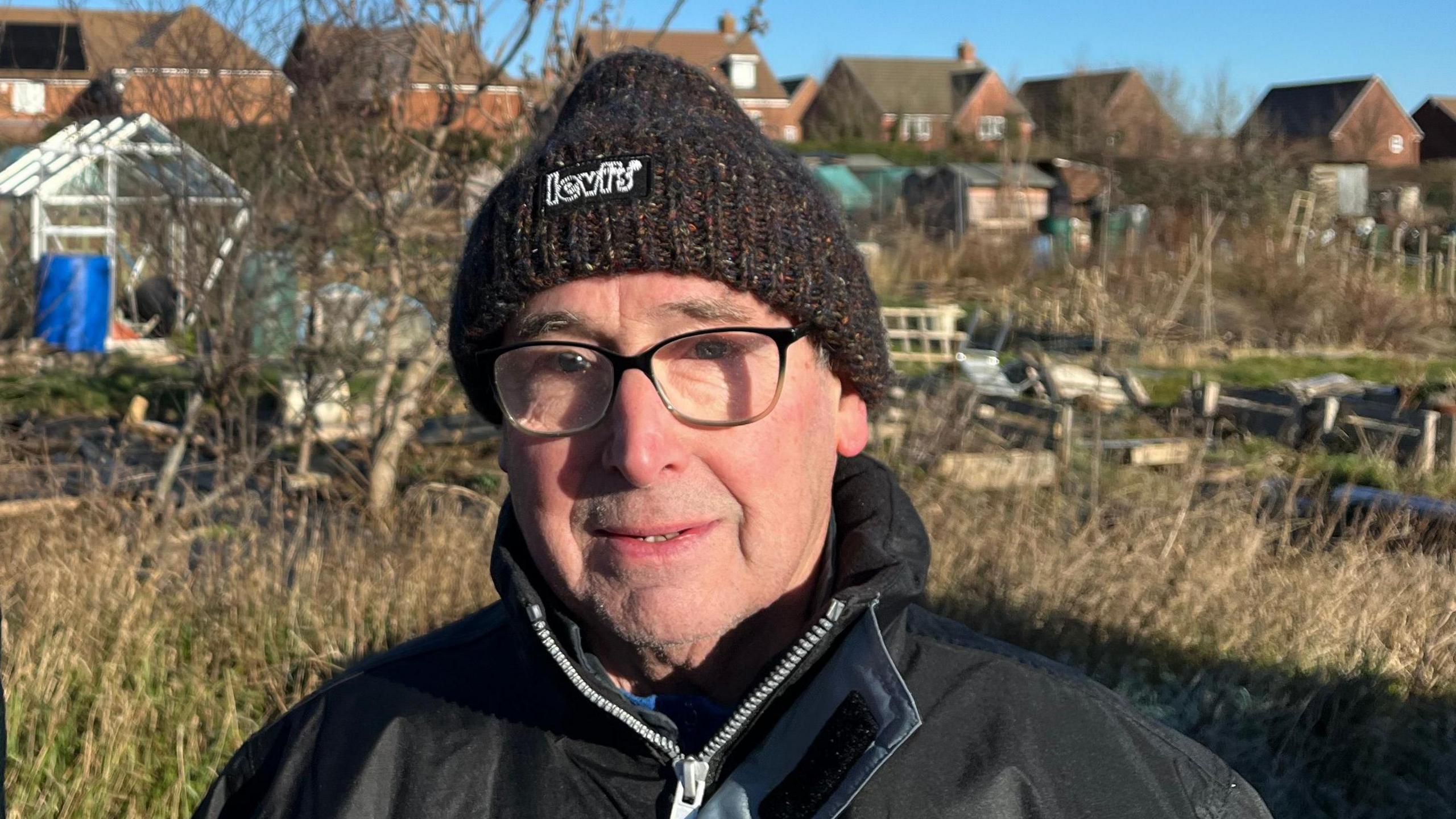 A man wearing a black winter hat and glasses. He is standing in an allotment with the backdrop of new build houses on a bright winter day.