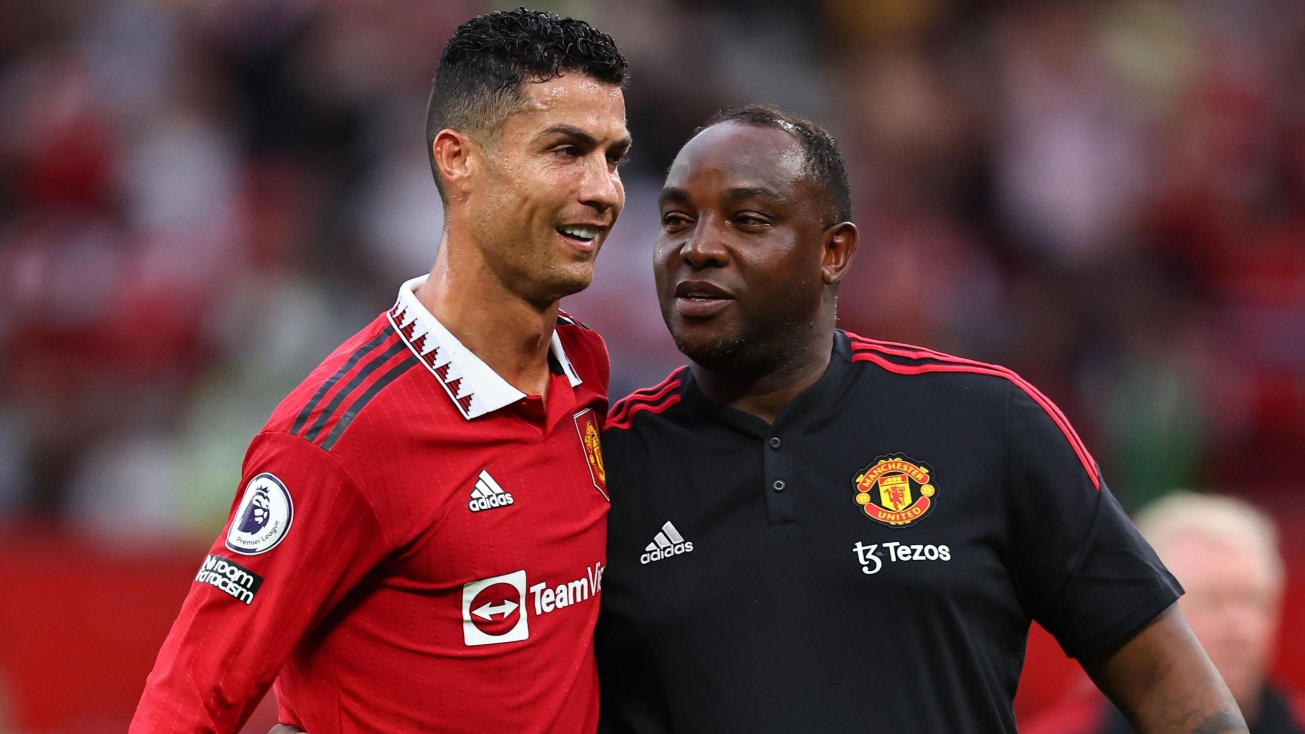 Benni McCarthy, wearing a black Manchester United polo shirt, smiling while holding his arm around the shoulder of Cristiano Ronaldo, who is wearing a red United home shirt.
