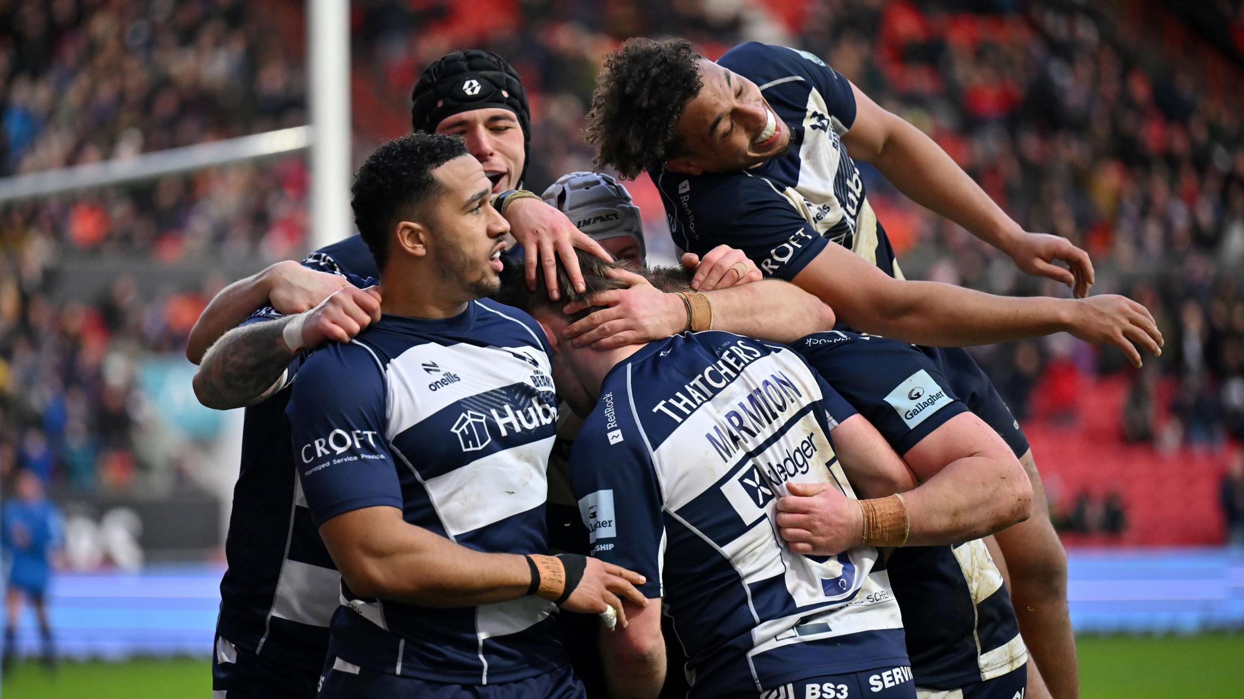 A group of Bristol Bears rugby players huddle together as they celebrate a try against Newcastle. A player is leaping in the air as he joins the group. The match is at Ashton Gate and the players are wearing Bristol's traditional blue and white hooped shirts