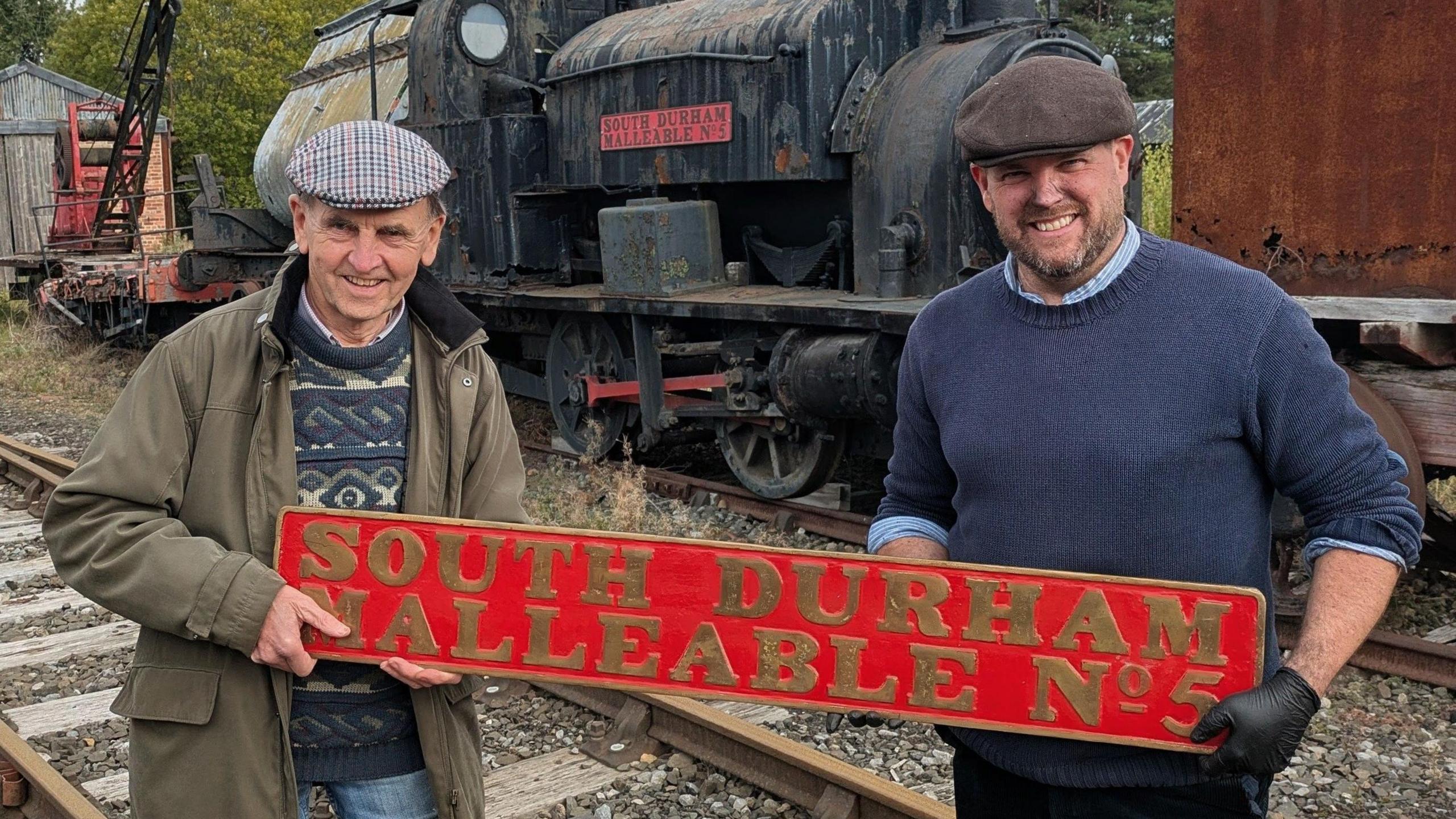 Tanfield Railway Trust chairman Derek Smith, left, and Beamish Museum director of transport Paul Jarman, holding between them an engine name plate - red with gold lettering reading South Durham Malleable No 5. Mr Smith is wearing a brown jacket over a sweater and Mr Jarman a blue jumper. Both are smiling and wearing flat caps. Behind them can be seen an aged steam locomotive in a state of disrepair.