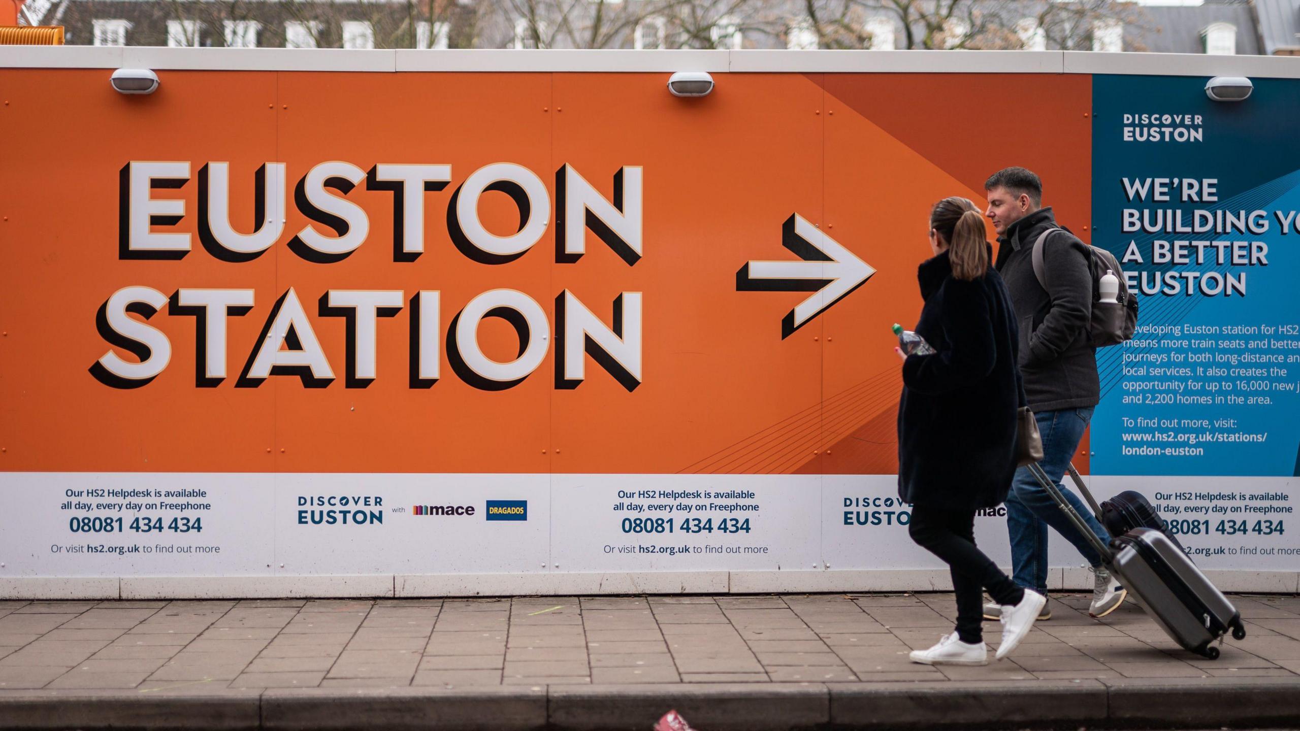 Two people walk with suitcases walk past construction hoarding at the HS2 construction site. The hoarding has information on it, including Euston Station written in large print with an arrow pointing commuters in its direction.