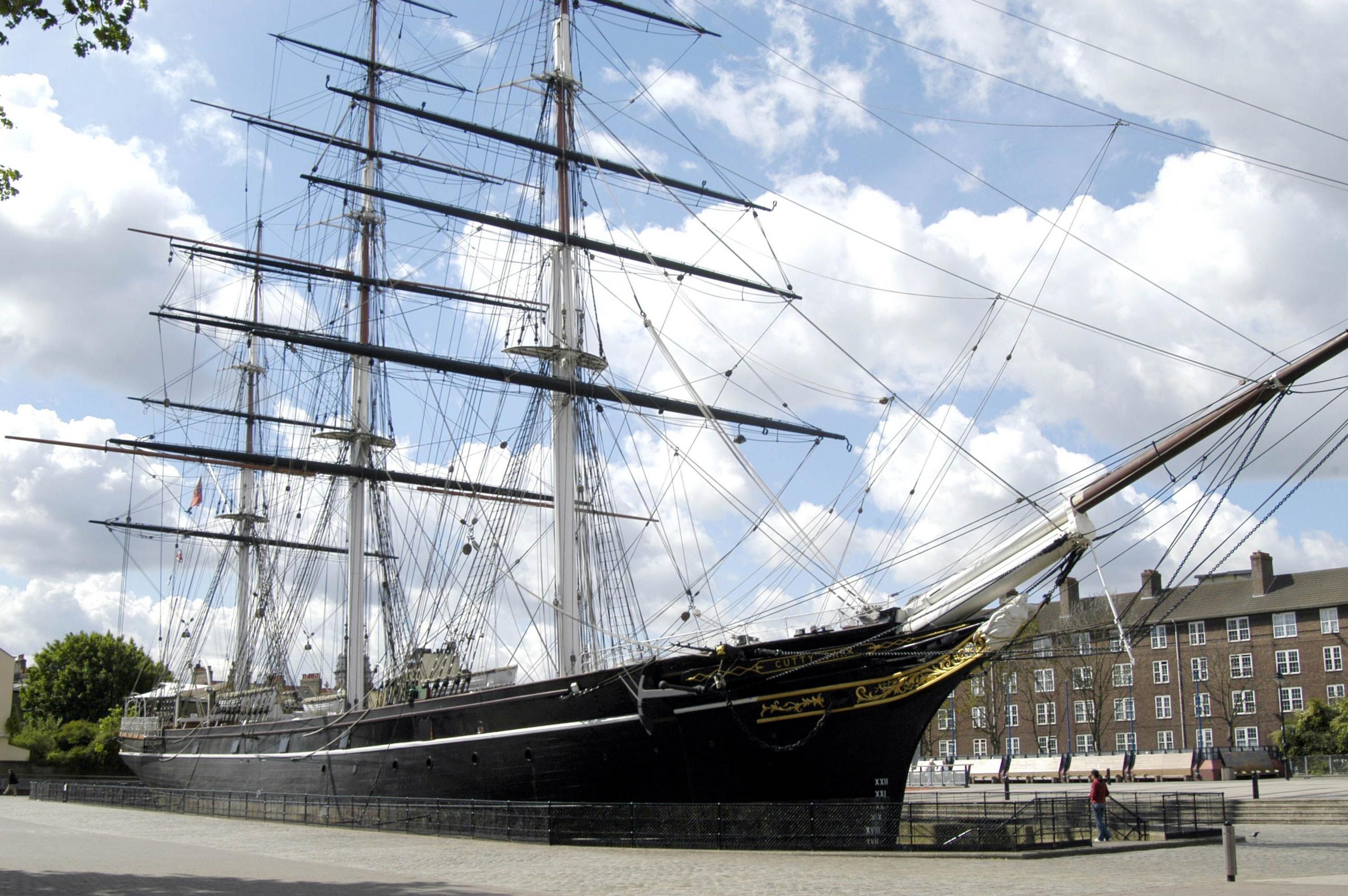 The 280 ft (85m) long, wooden boat has three masts. The body of the ship is painted blue and gold. It is moored in a fence-off area surrounded by pavement. A block of flats can be seen in the distance.