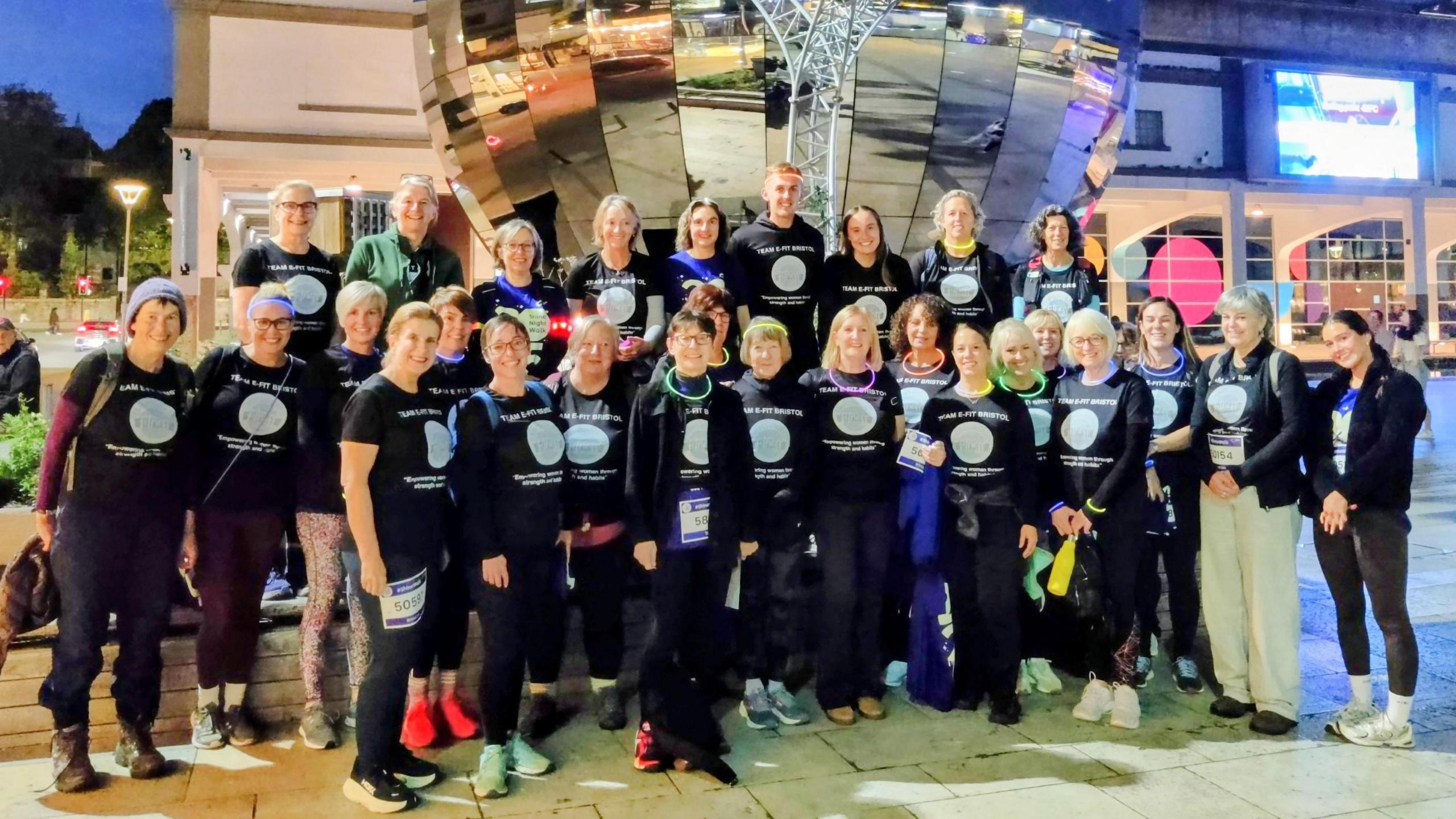 A large group of people wearing dark T-shirts and trousers standing in front of a large silver globe in Millennium Square in Bristol. The picture is taken after dark and the area is lit by streetlights