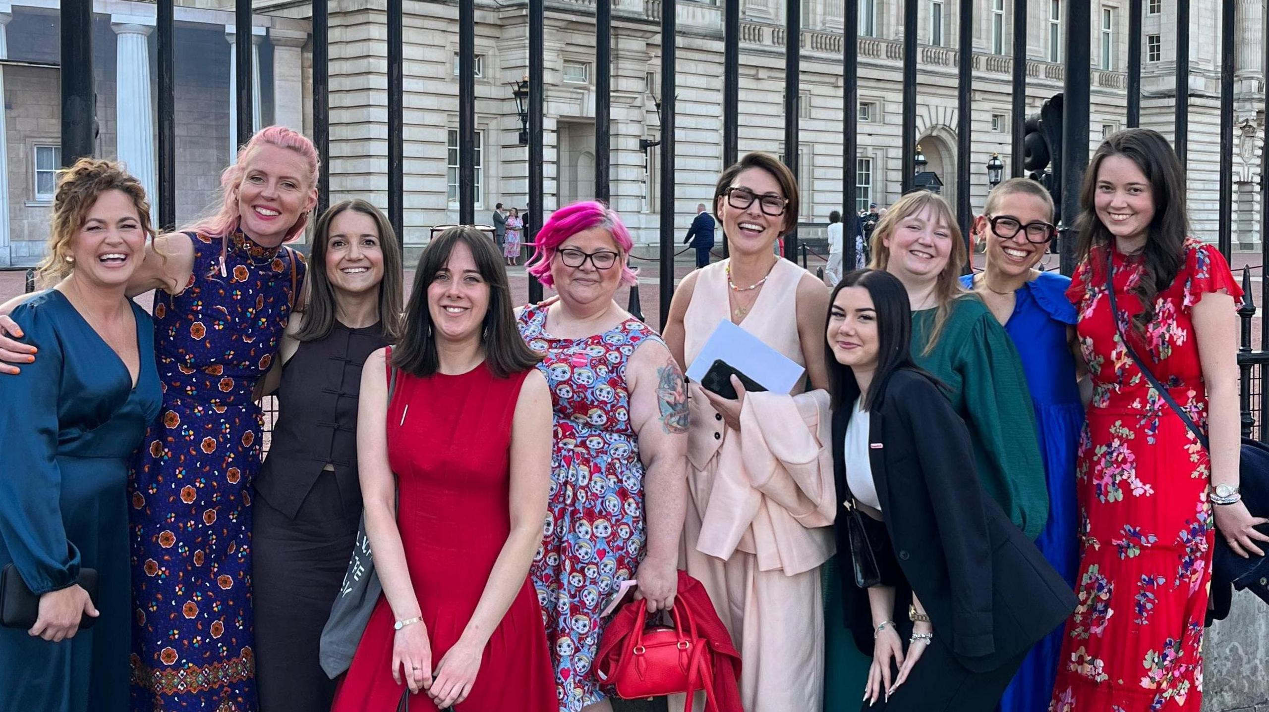 A group of women standing together and smiling at the camera outside Buckingham Palace.
