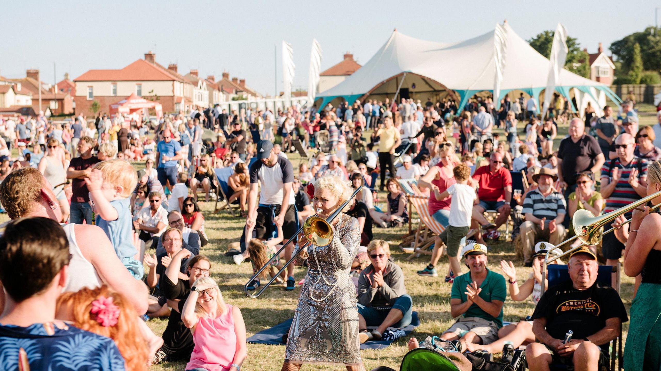 A woman in a silver dress playing a trombone amongst a crowd of festival-goers