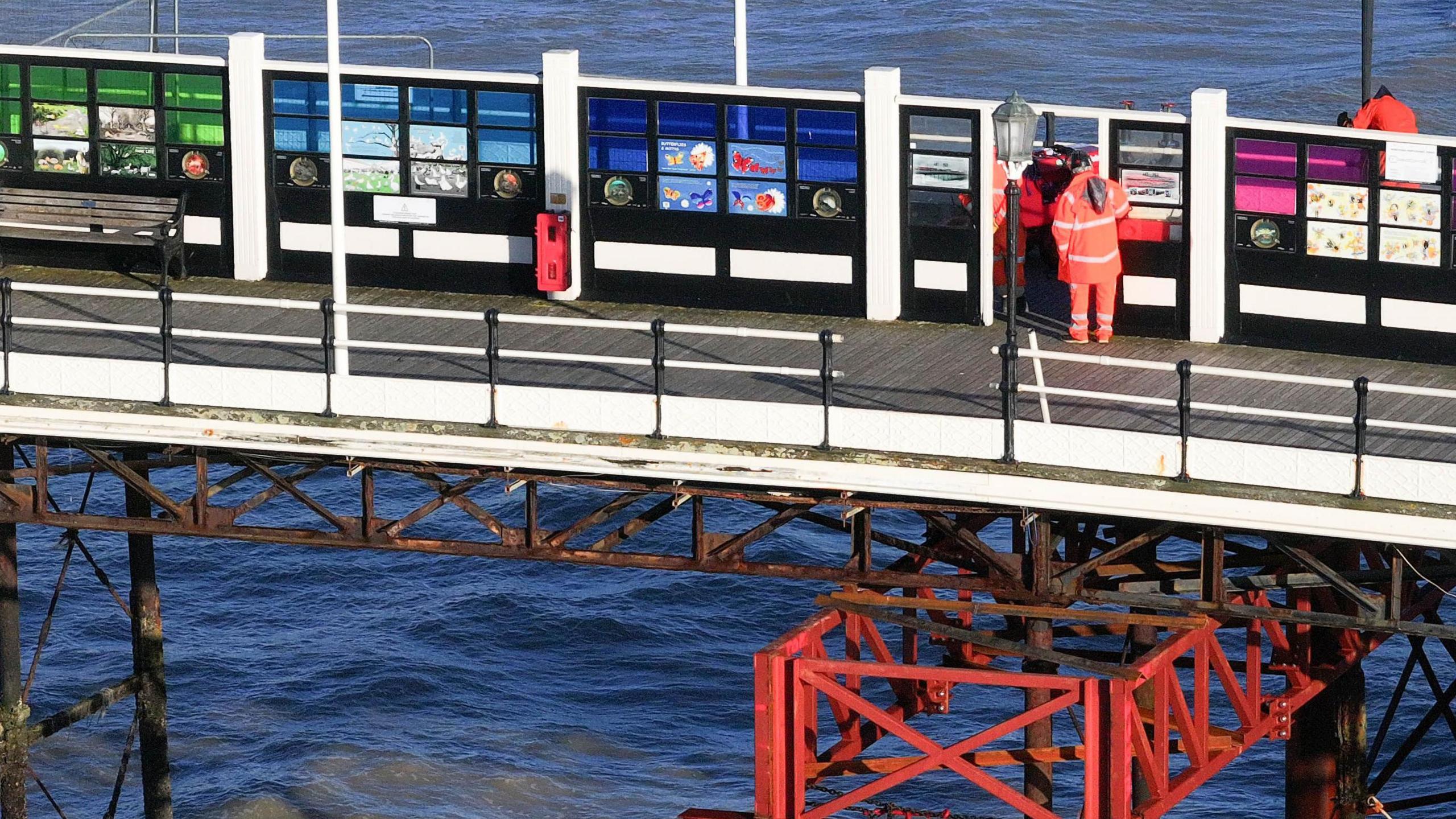 Men in orange hi-vis work on the pier while underneath there is orange scaffolding on the supports. 