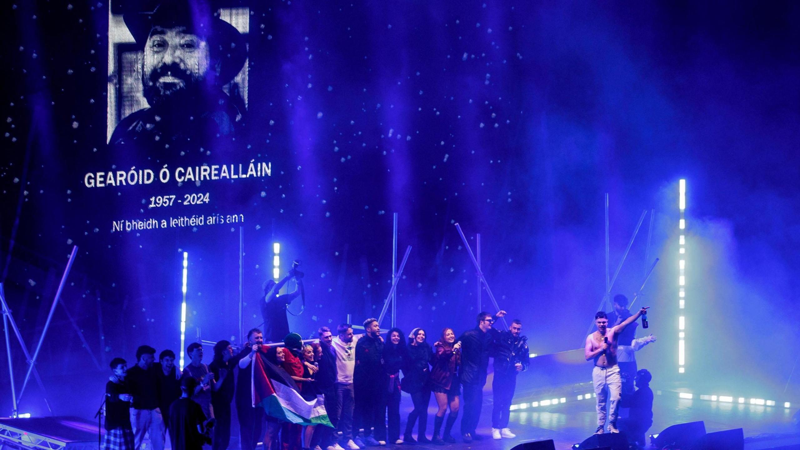 About 20 people are lined up shoulder to shoulder on a stage - to the right is a member of the rap group Kneecap who is shirtless and holding a microphone. The group are holding a Palestinian flag. On a screen in the background is a picture of Gearóid Ó Cairealláin with his name, date of birth and year of death and the Irish saying which means: 'We will never see his like again.'