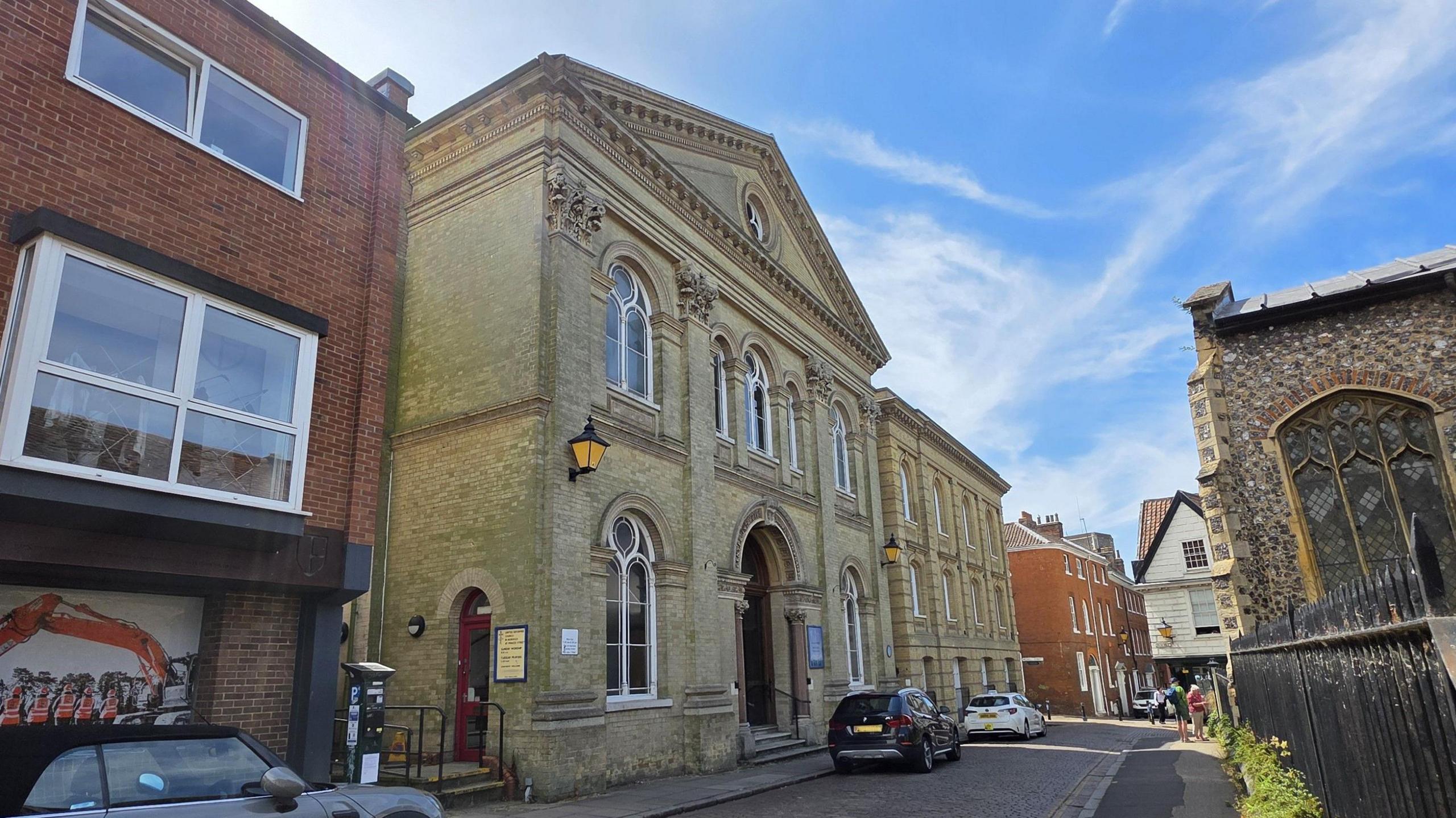 We can the 19th century church on a road in Norwich. Across the street we can see another church - and three cars are parked on the road. Above, is a bright blue sky with light cloud.