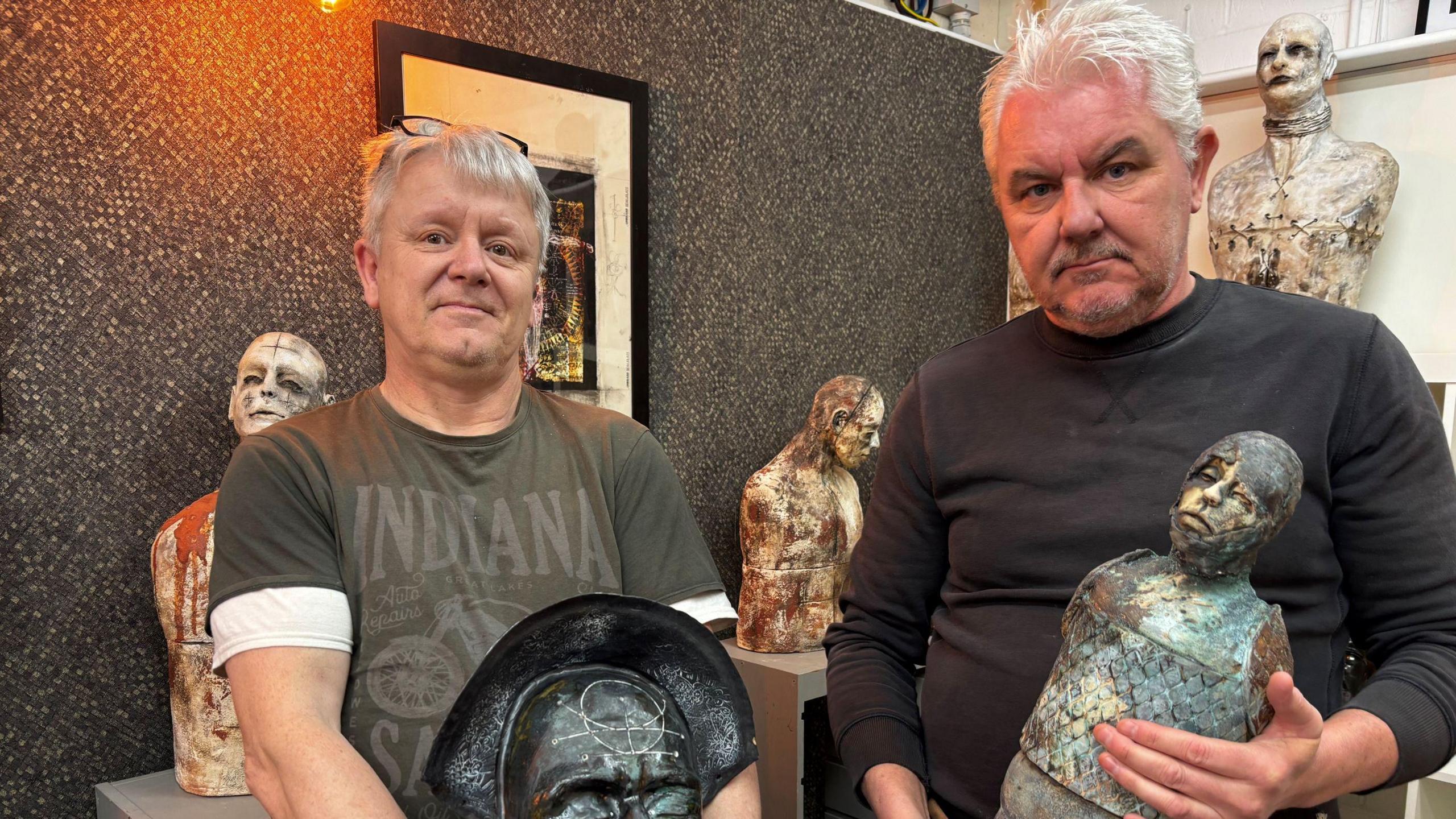 Paul and Jason Skellett stood up next to each other holding sculptures. Behind them are sculptures on display. 