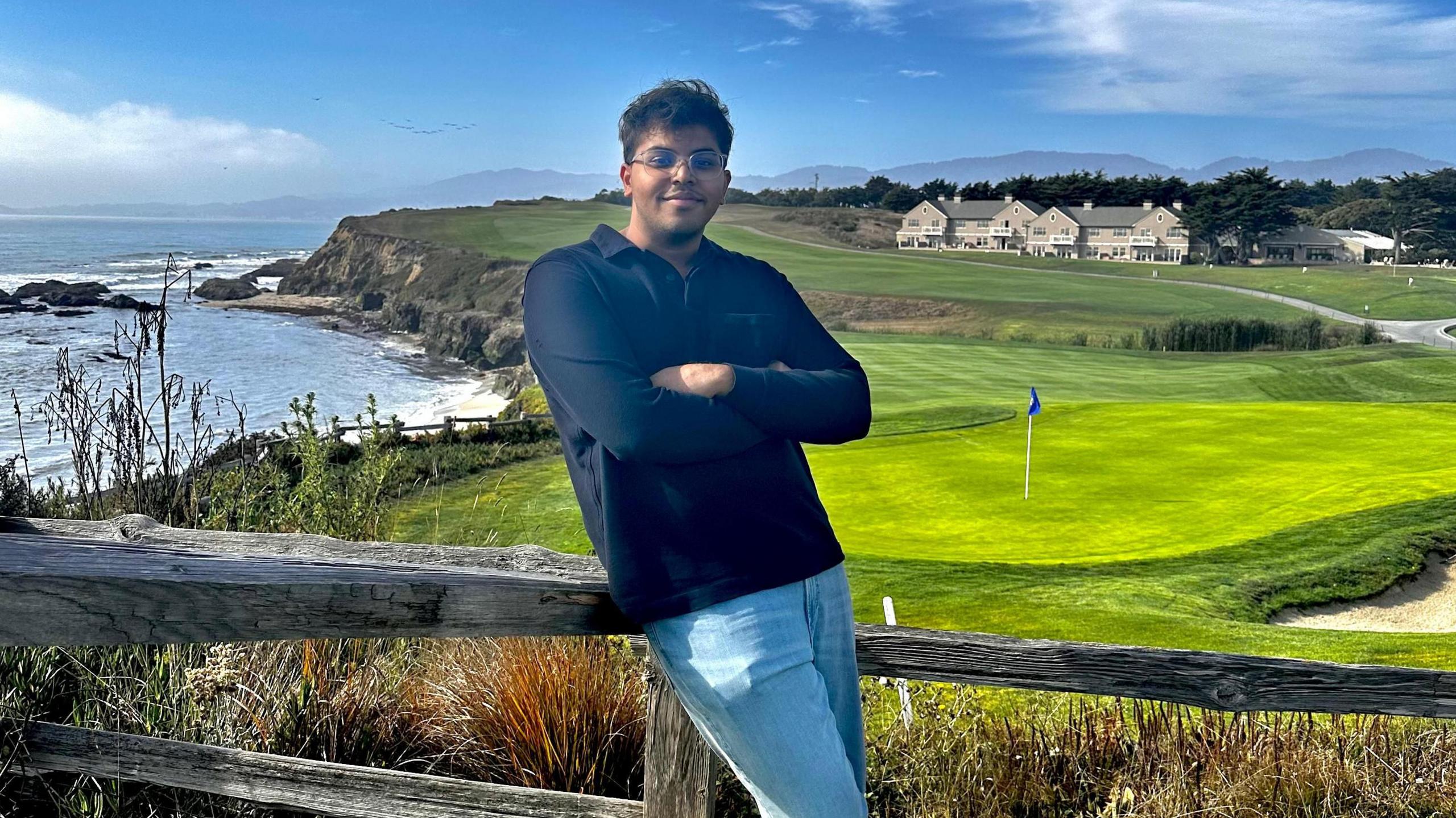 Ayush Deb poses for a picture with his arms crossed in front of a golf course by the sea.