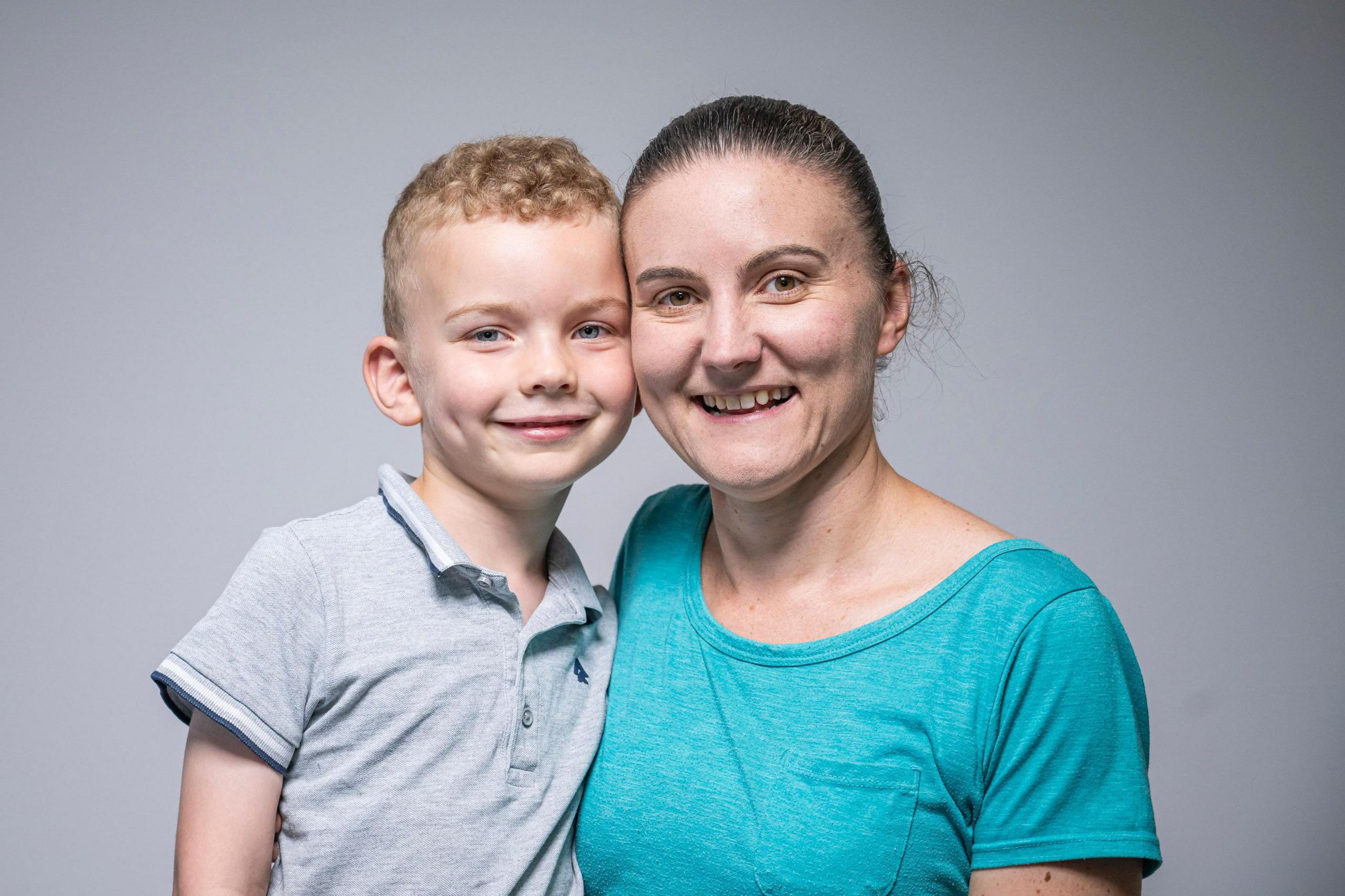 A portrait photo of Aiden and mum Steph. He has short blonde hair and wears a grey collared short-sleeved shirt. Steph has dark hair tied back, and a blue t-shirt. They are smiling, their cheeks touching.
