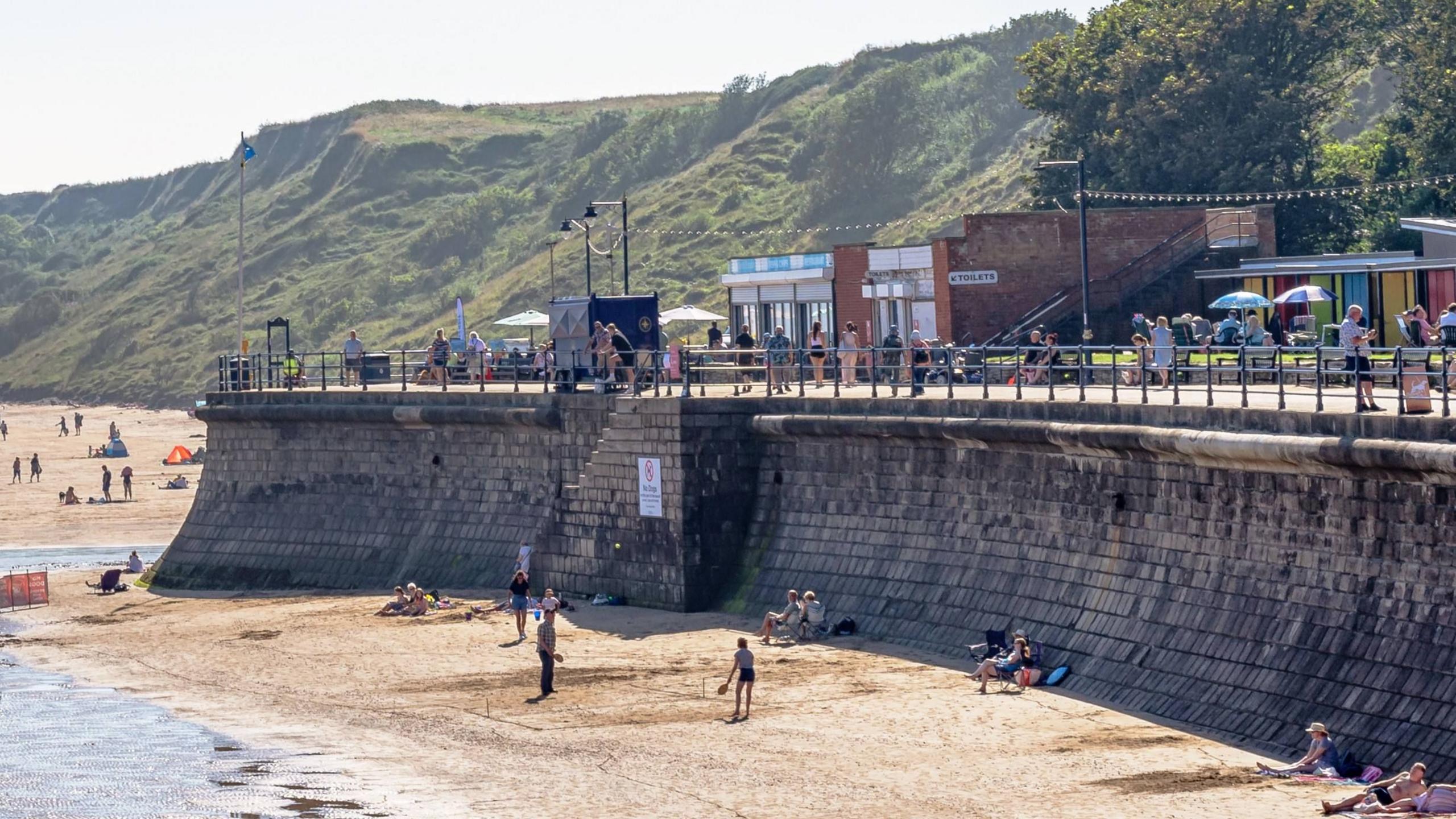 A small block with a sign reading "toilets" which is next to a fish and chip shop. It is above a beach with a hill behind it, and people are walking along the promenade in front of it.