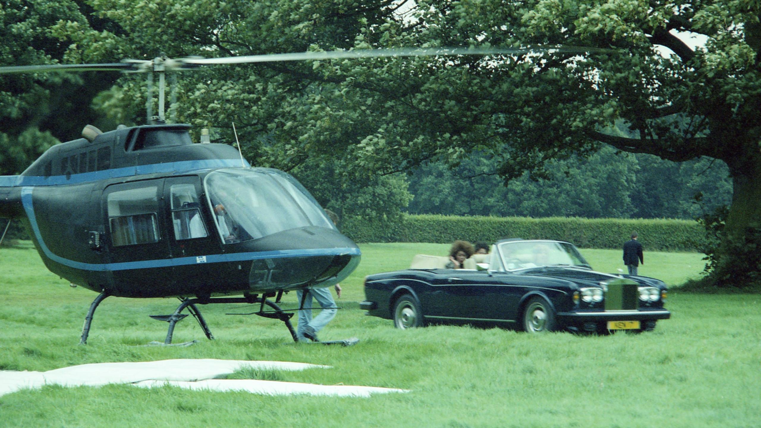 A colour shot of a green field with trees in the background in daylight, with to the left of frame a black helicopter with its rotors moving having either just landed or preparing for take-off. To the right of frame is an open-topped Rolls-Royce, with three women being driven in it as passengers. Next to the helicopter slightly to the foreground appears to be a large white 'x' shape marked out with sheets or boards.