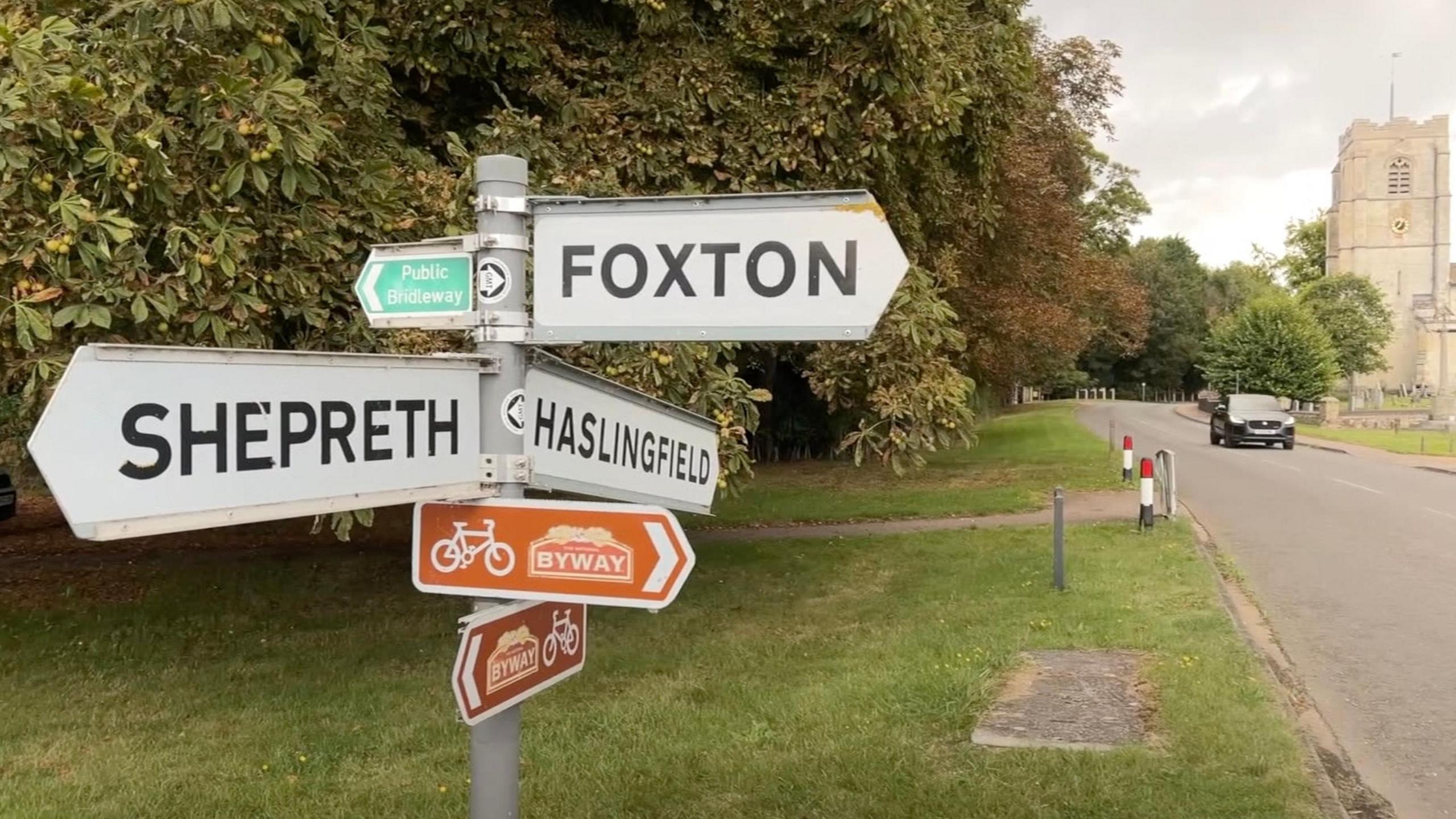 Signpost showing affected villages in South Cambridgeshire next to a tree-lined road with a car and church in the background.