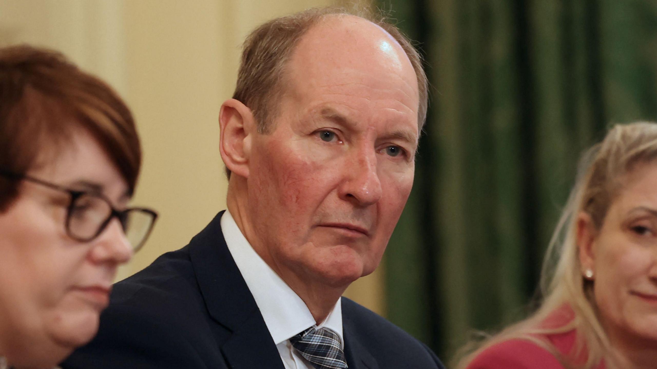 Alan Armstrong, CEO of Almac, is sitting at a table with other people. There are women sitting next to him. He's wearing a black suit with a shite shirt and a patterned tie.