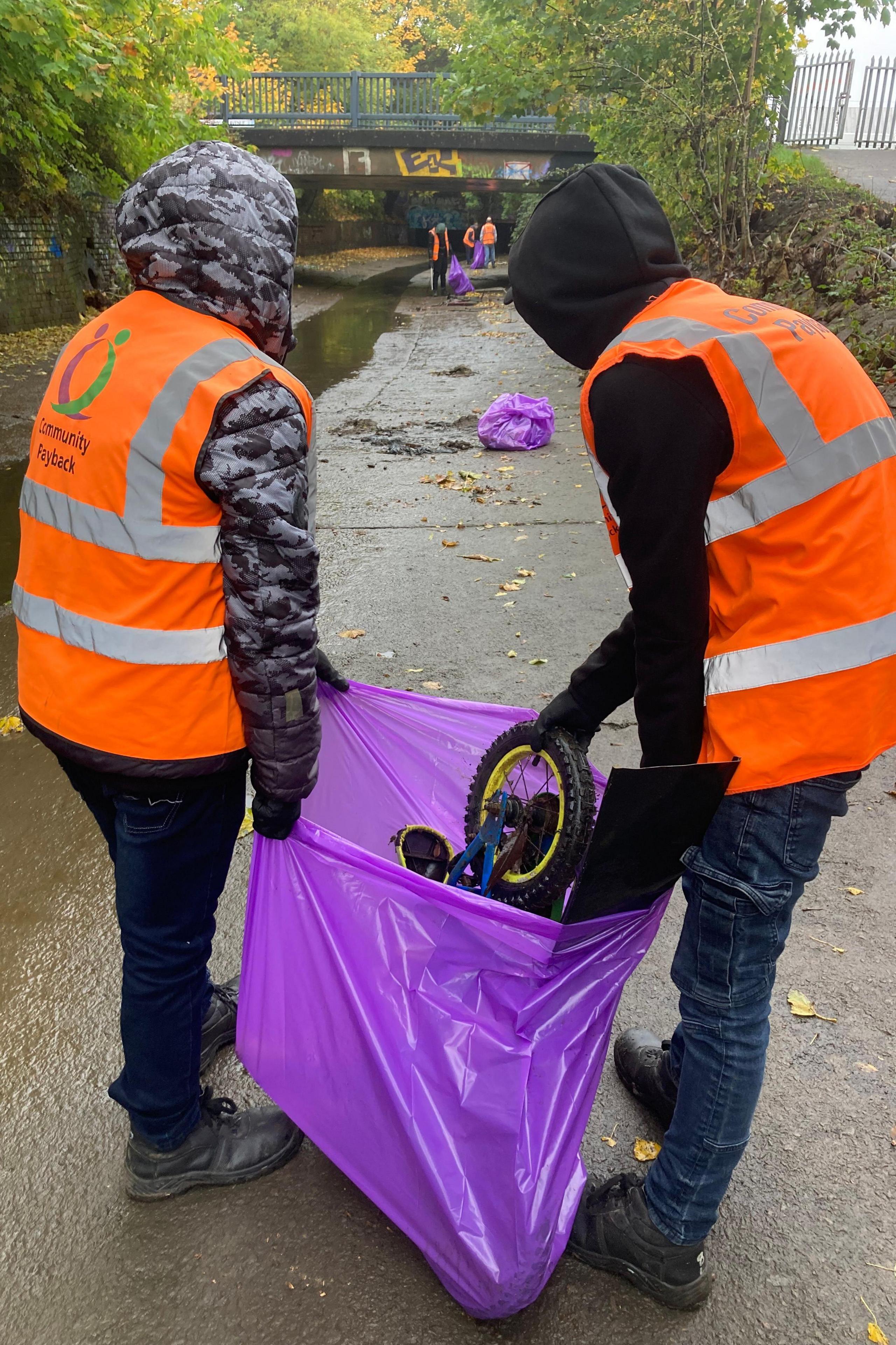 Two people in hi-viz jackets are carrying a bag full of waste items that have been thrown into Willow Brook in Leicester. They are offenders carrying out community service. 