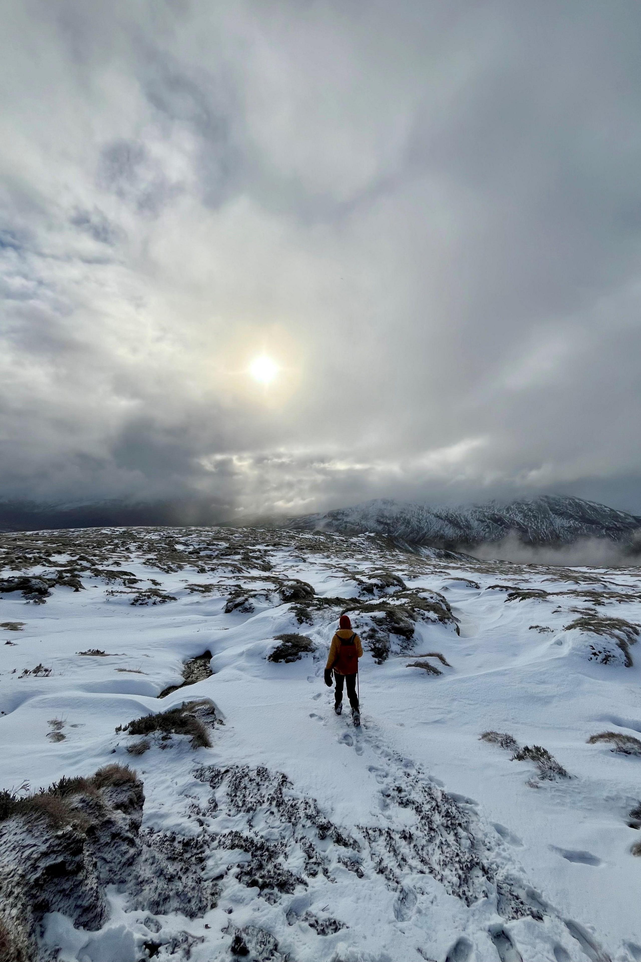 Hiker on snow-capped hill, with peaks in distance and sun shining through clouds. 