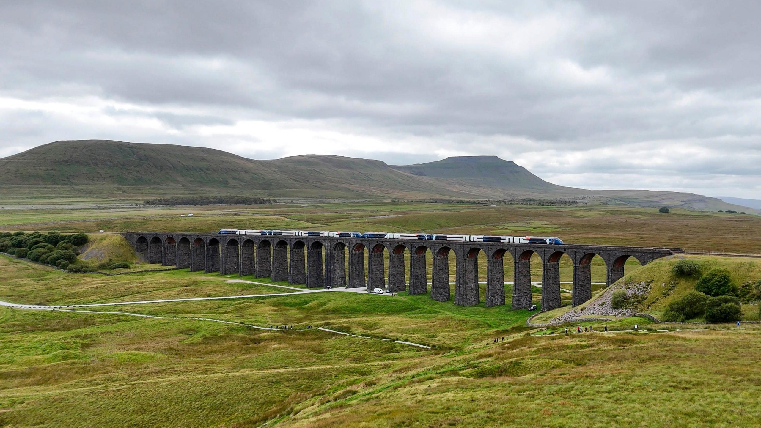 A train running along the Settle to Carlisle railway line over the Ribblehead Viaduct. The stone viaduct with its many arches is surrounded by rolling green land with fells in the background.