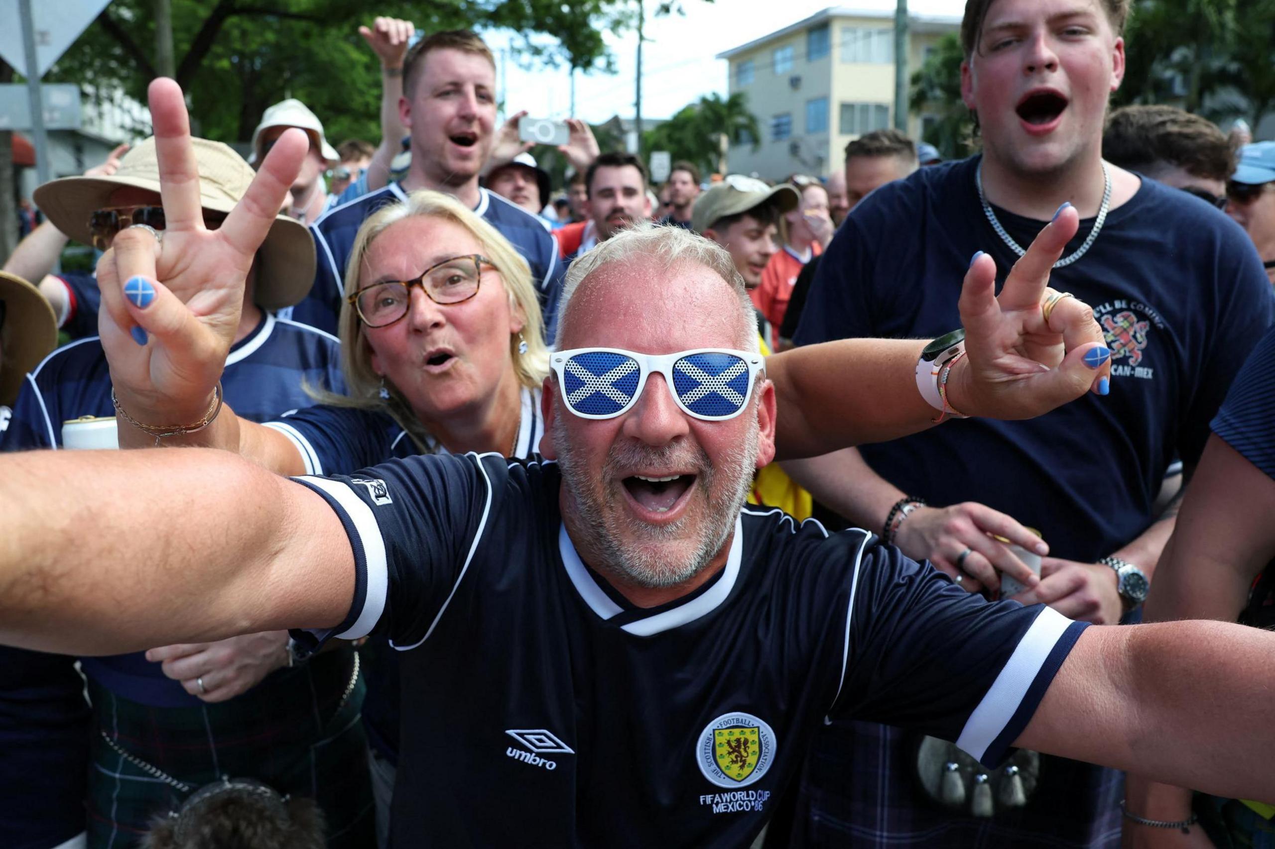 Scotland fans in Miami. A man wearing sunglasses with Saltires on them, and wearing a dark blue Scotland top smiles as he reaches out as if to hug the photographer. Other Scotland fans stand behind him.