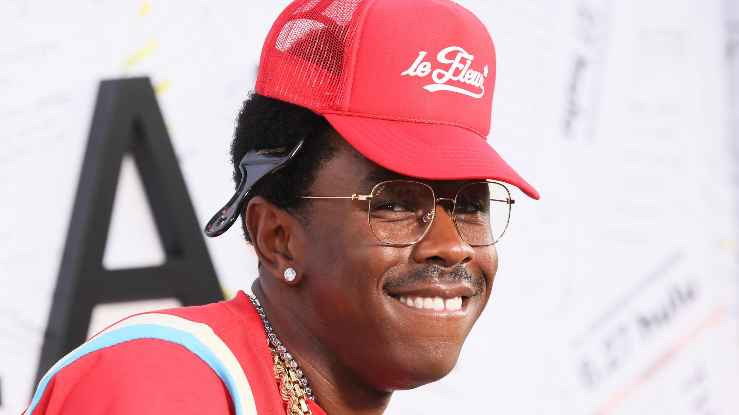 Tyler, The Creator wearing a red cap and glasses, smiling.