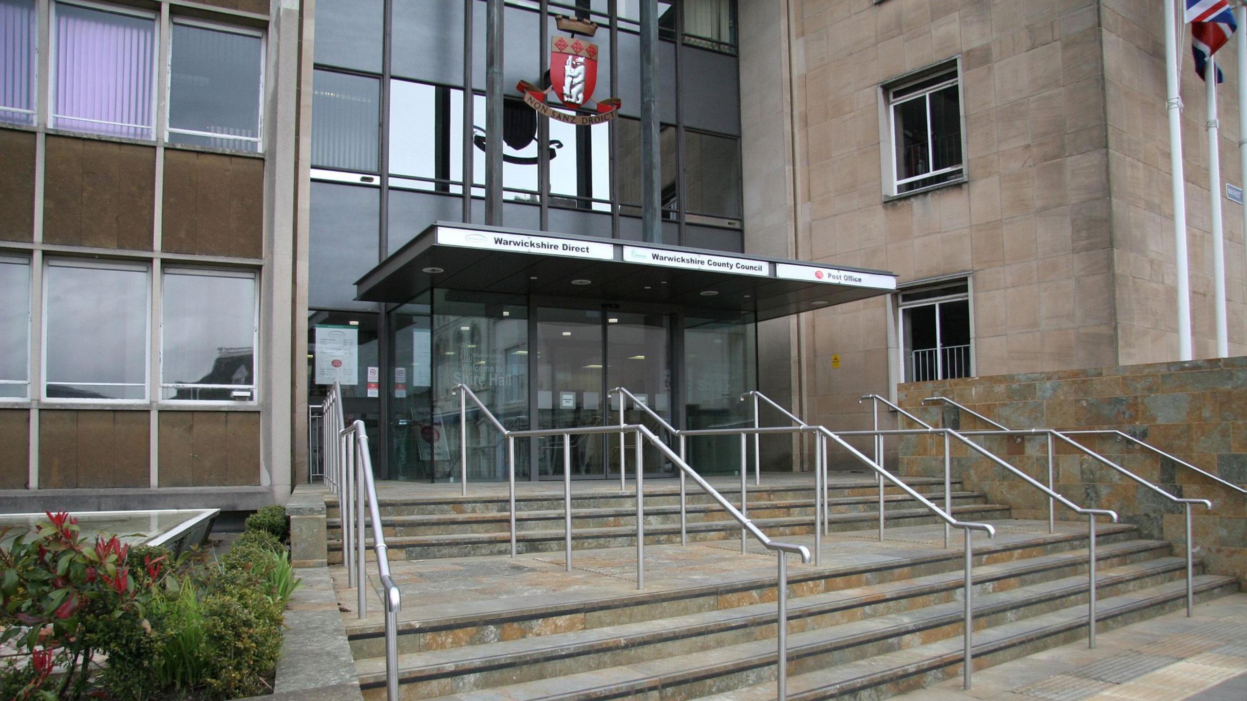 The outside of Shire hall, a stone and glass building with a canopy over the front door topped with a red and white shield. A union jack is flying to the right of the door.