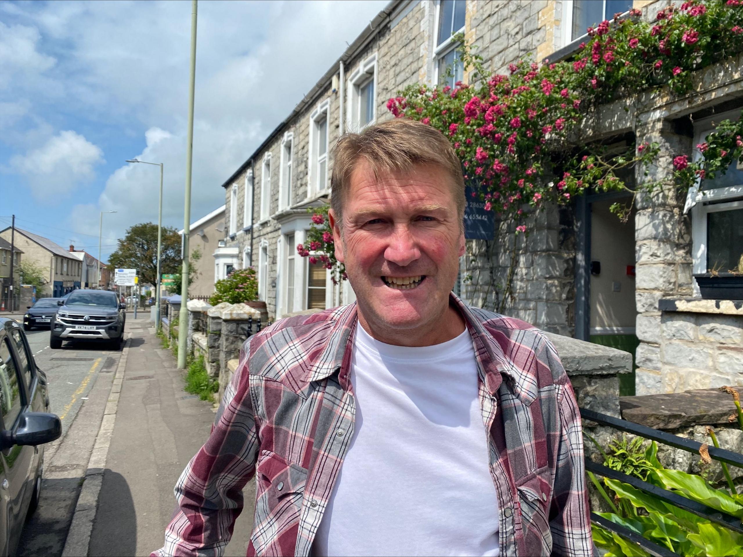 Llun o Huw Tudur yn sefyll ar balmant ar ddiwrnod braf.