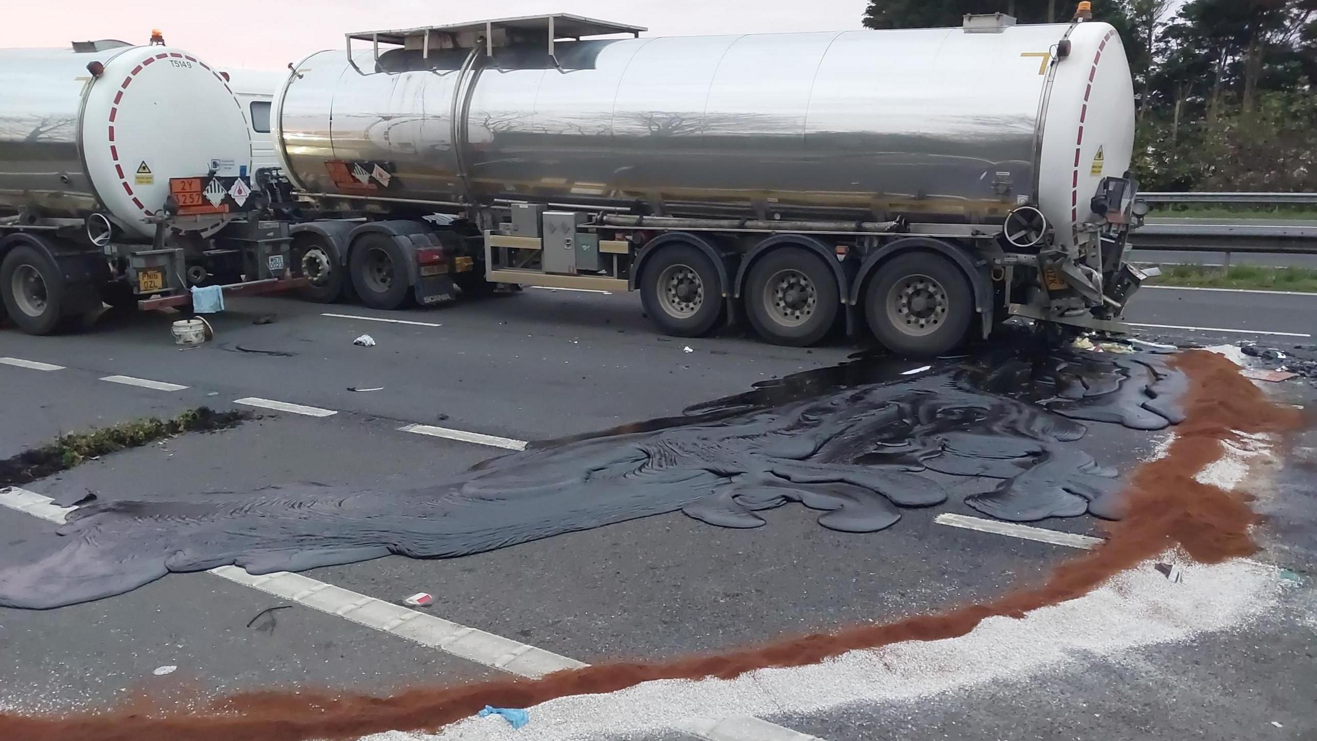 Tanker with black liquid spilling on to a motorway - and another alongside it - an edge of white and brown powder surrounds the spill