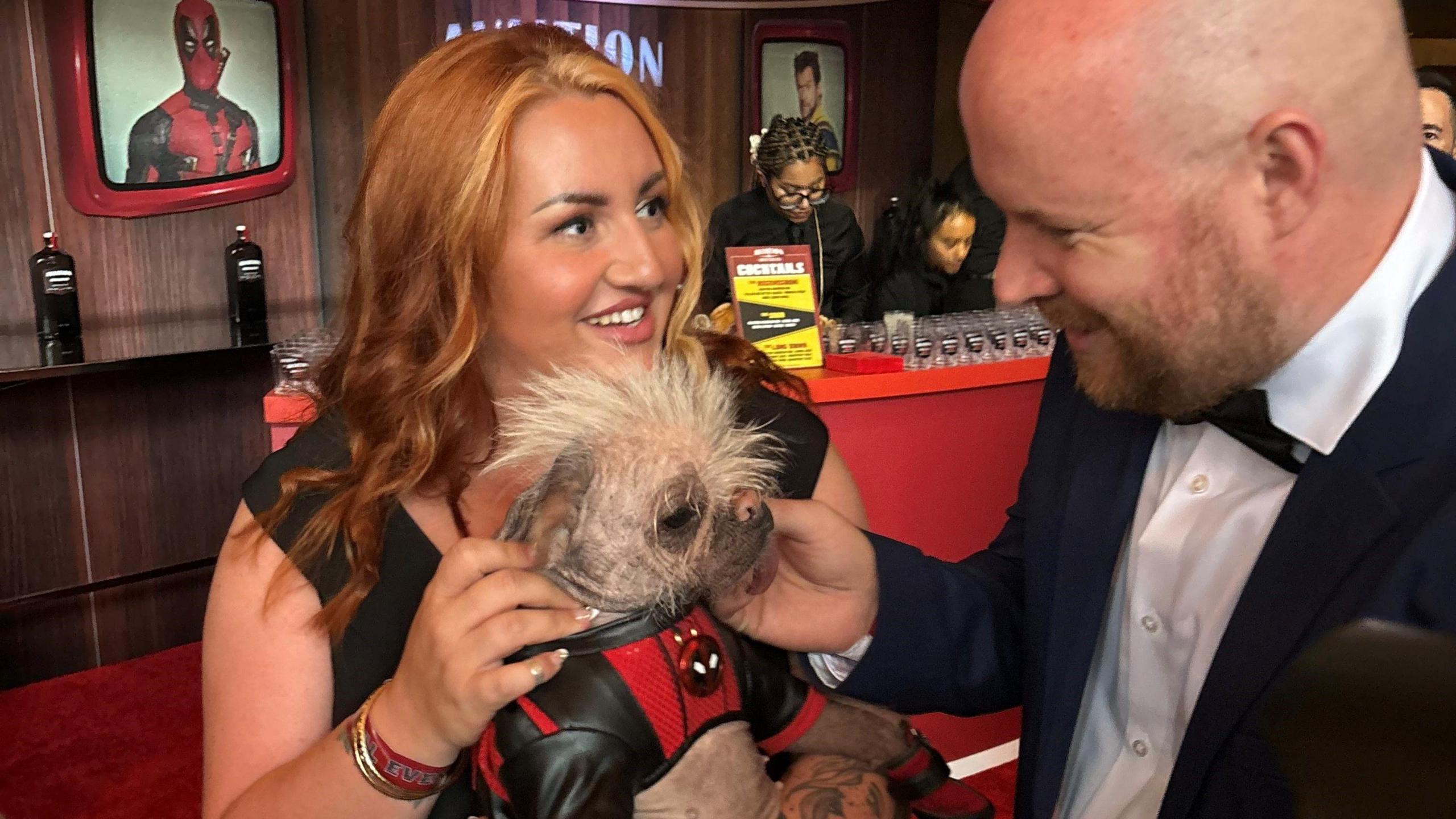 A smiling Holly Middleton, who wears a black dress, holds her dog Peggy, who is wearing a black and red superhero costume, while her husband Luke, who wears a dinner jacket and white shirt, tickets Peggy's chin.