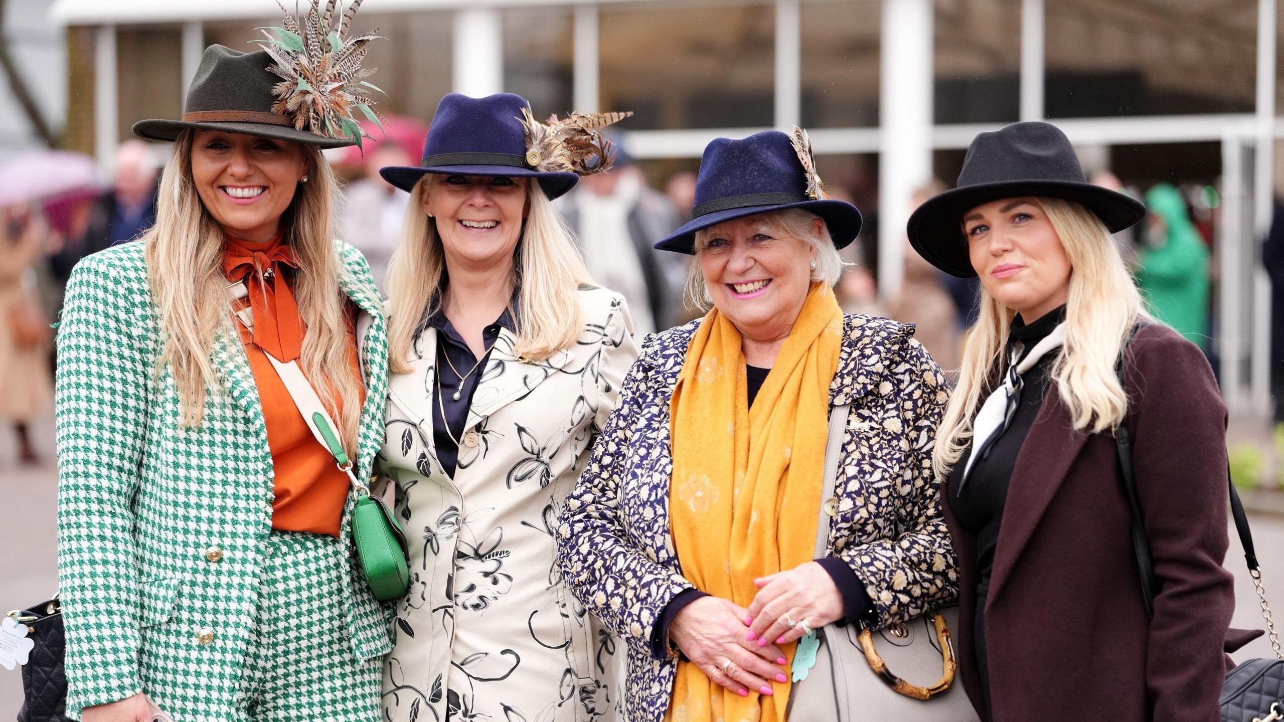 Four women in smart outfits stand in a line and smile for the camera at the start of day three of the Cheltenham Festival. All four have long blonde hair and are wearing hats