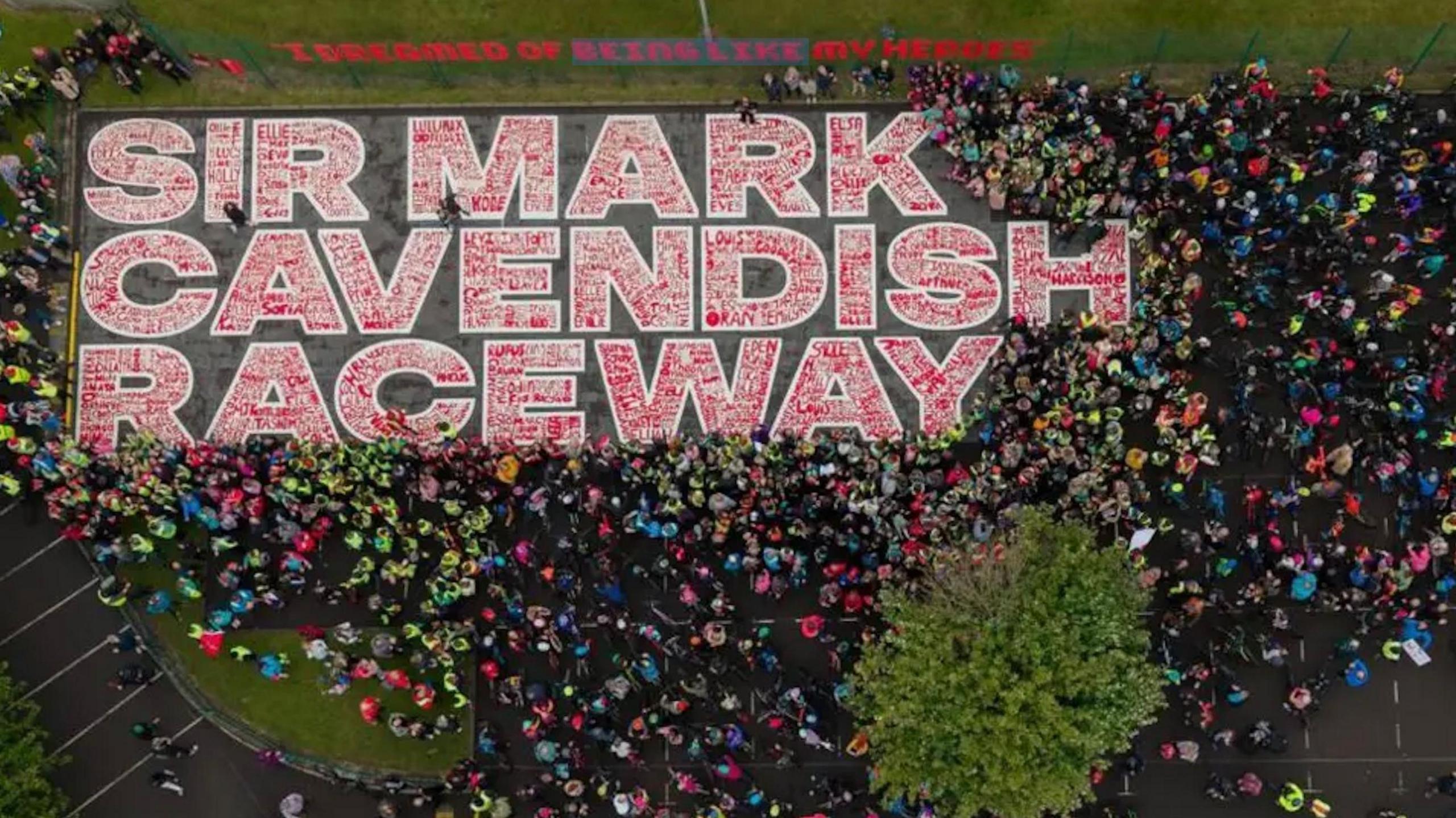 The mural, from above, is outlined in white with the names of children in red. There are hundreds of children standing around wearing bright colours.