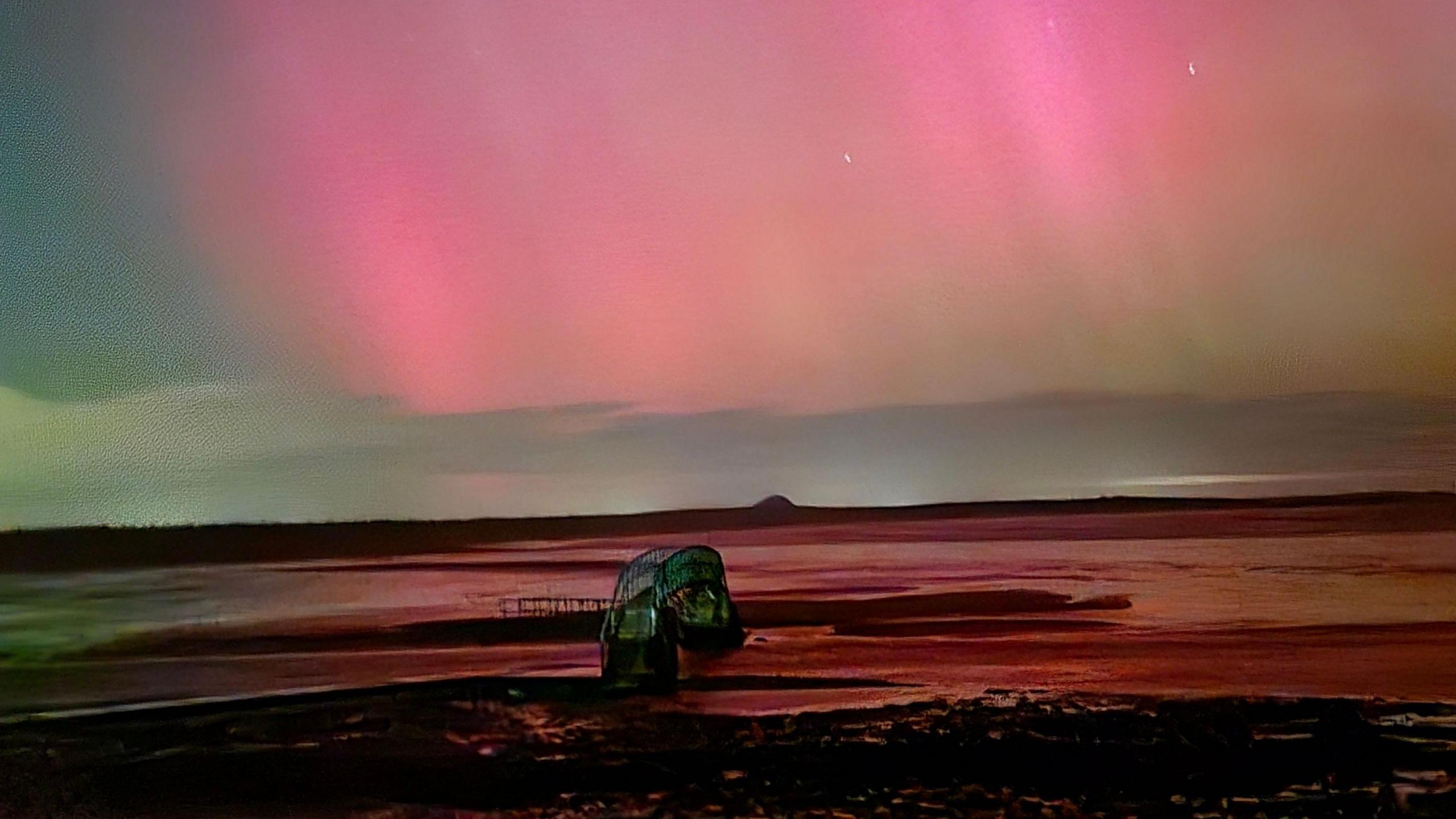 The bridge at Belhaven in East Lothian crossing a waterway. The landscape is almost entirely flat but for the hump of Berwick Law in the far distance. Red and pink aurora touch the clouds on the horizon.