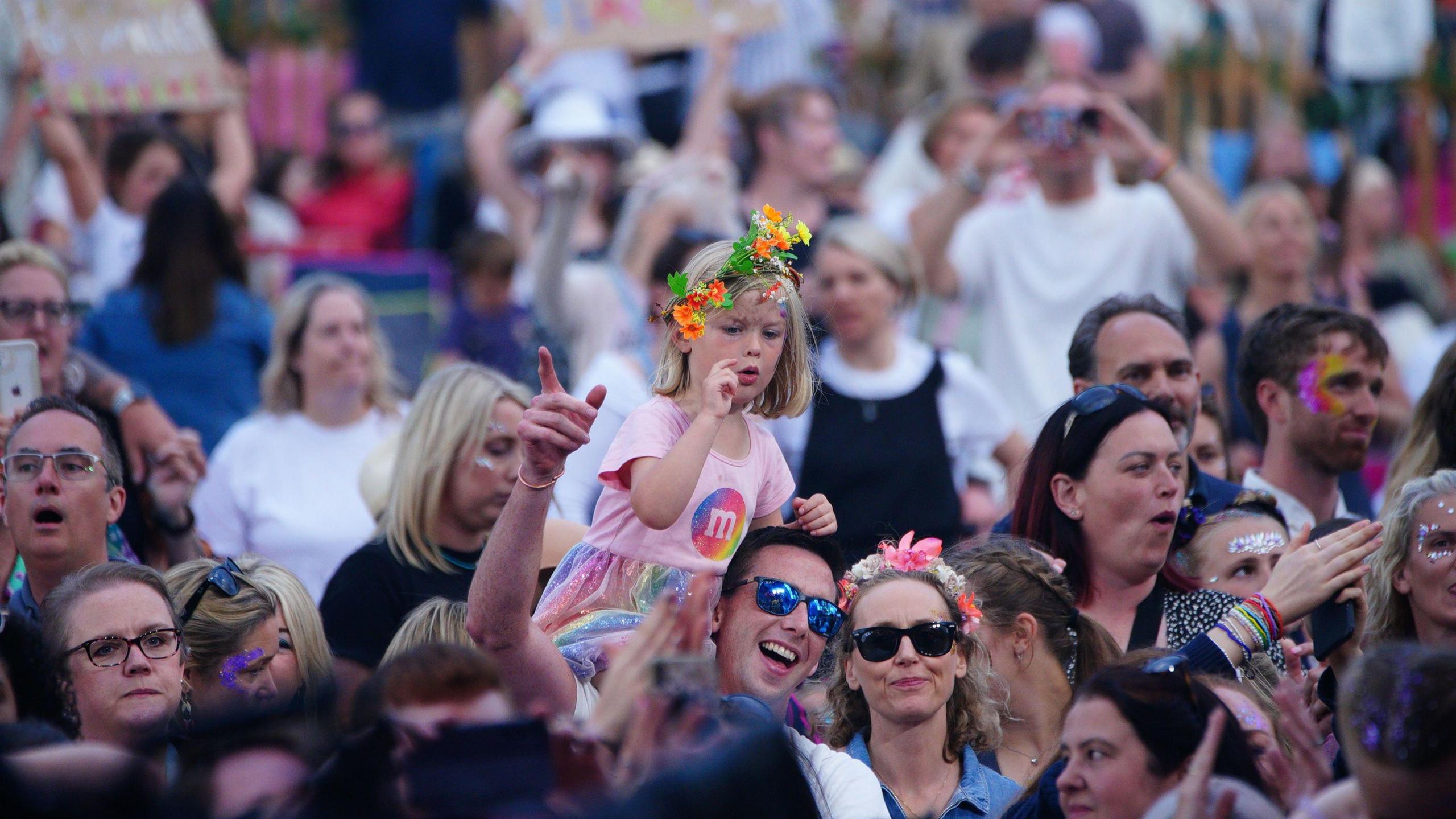 An image of the crowd at Flackstock. A young girl in a bright pink t-shirt and skirt. She is sat on the shoulders of a man.