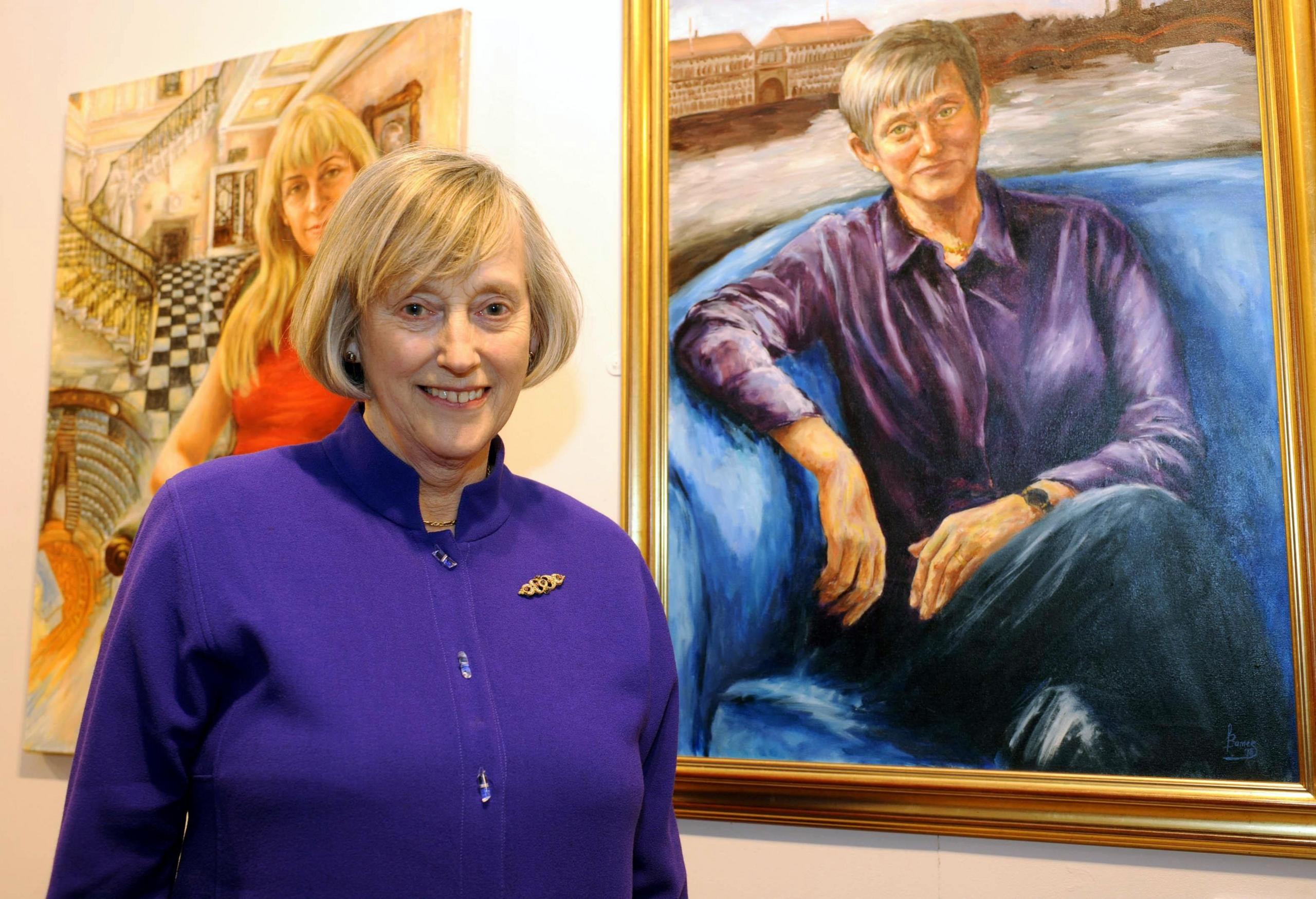 Dame Stella Rimington smiling by a portrait of her. She is looking at the camera and wearing a purple top, which echoes the purple shirt in the portrait 