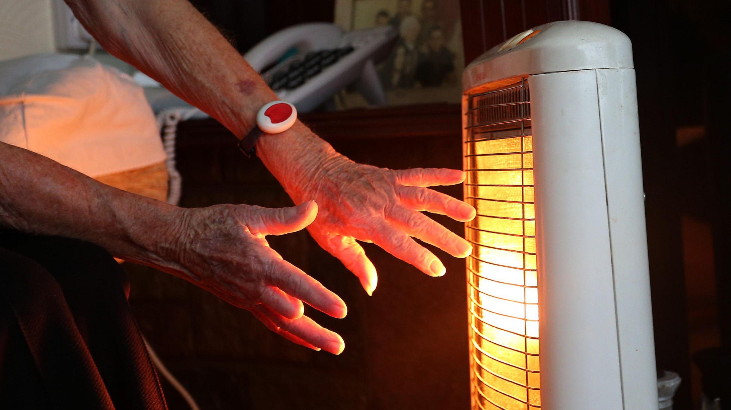 An elderly person warms their hands in front of an electric fire