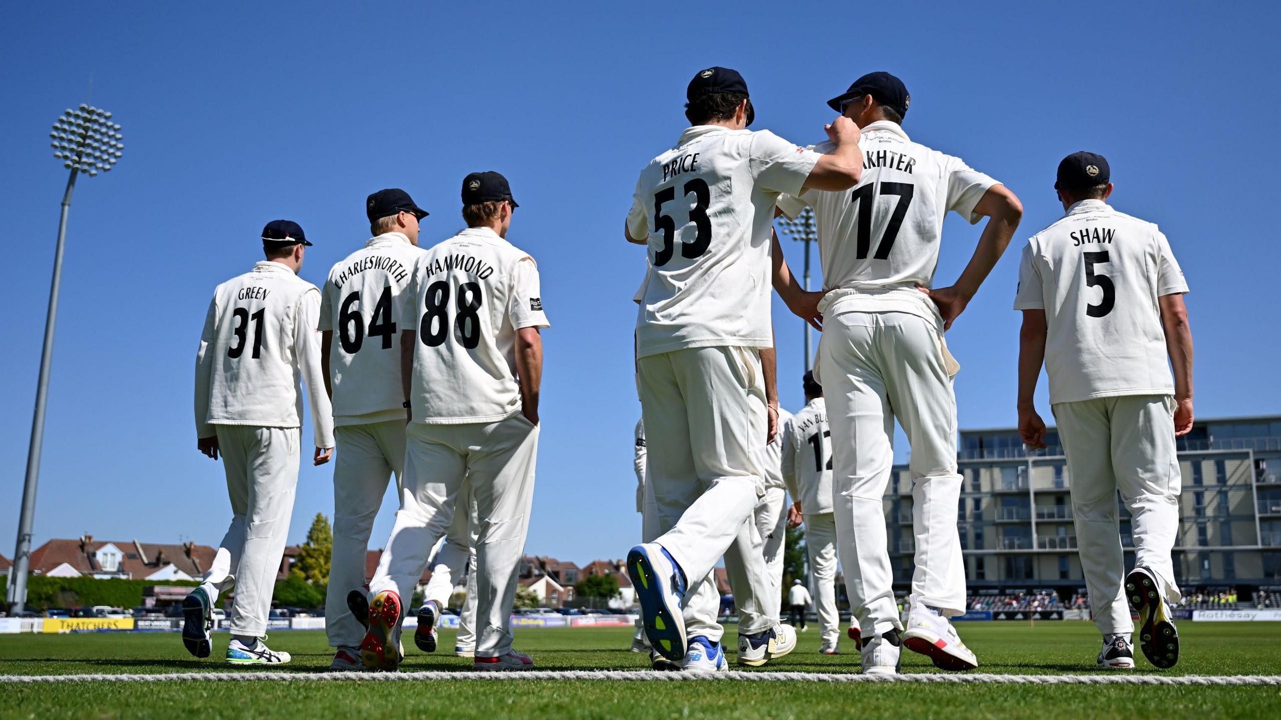 Gloucestershire's cricketers walk over the boundary rope onto the field ahead of their match with Kent at the County Ground in Bristol. In the background one of the stadium floodlights is visible as is the block of flats that overlook the pitch