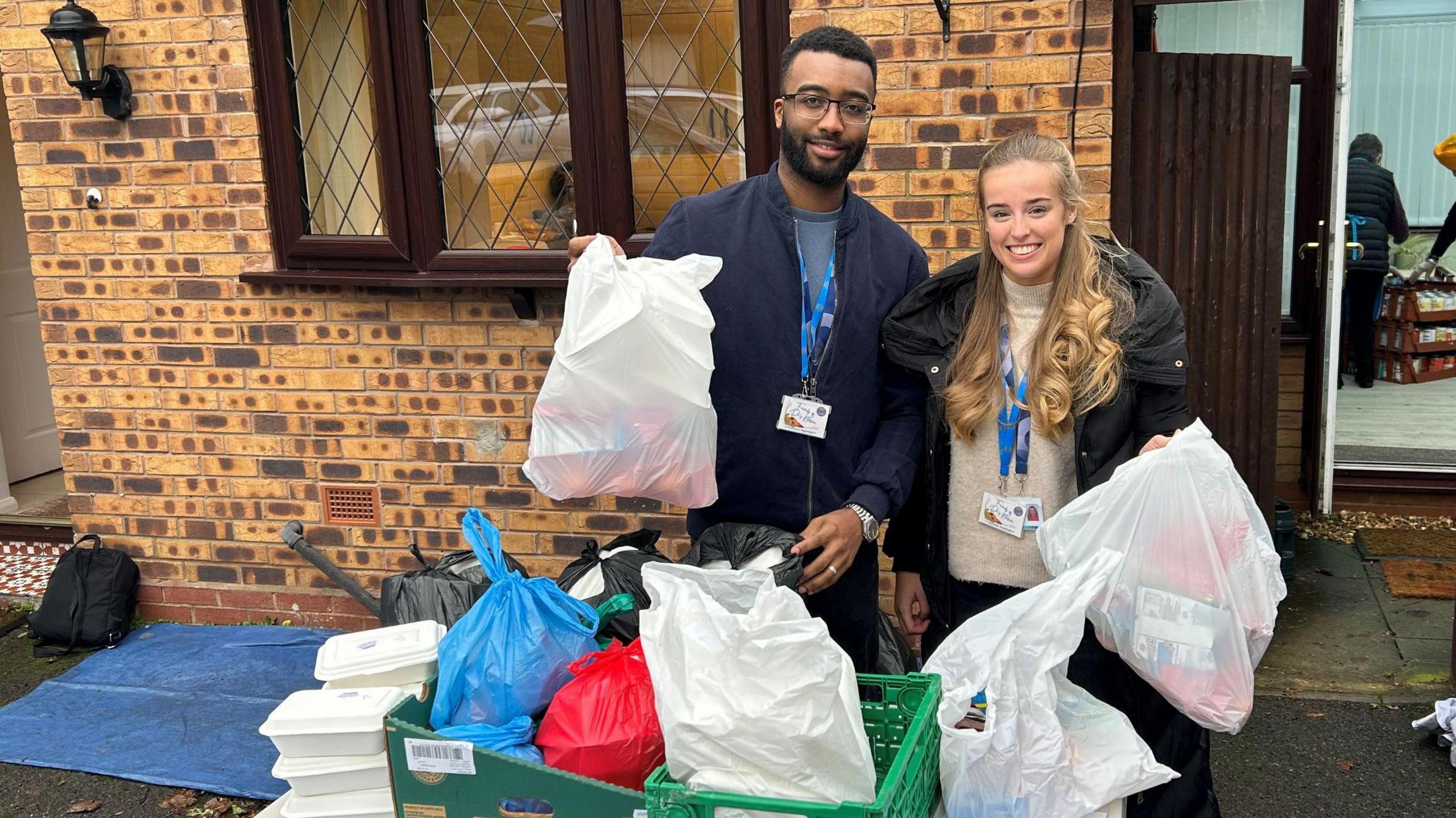 Volunteers pictured with bags of food. A man wearing a blue jacket and glasses holds up a bag of food items. A woman next to him has long blonde hair, a beige jumper and black coat. Both wear work identification and are smiling at the camera. 