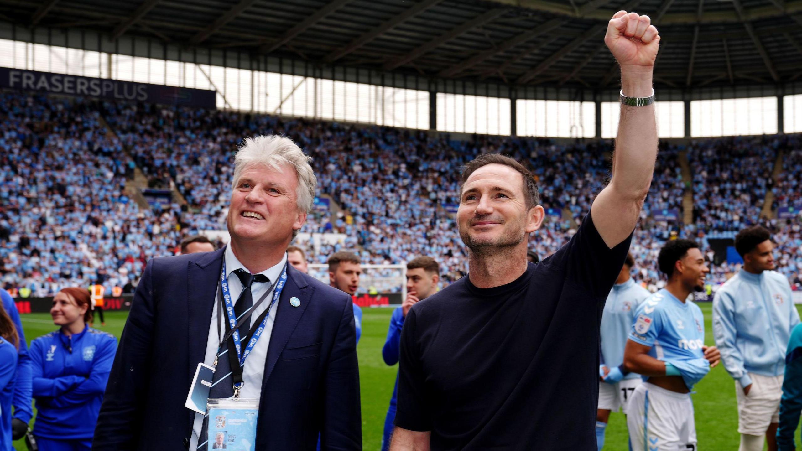 Doug King (left) has white hair and is earing a blue suit, white shirt and blue tie with two lanyards around his neck. He is stood next to Frank Lampard, Coventry City's head coach. Frank is wearing a black T-shirt and has one arm waving up at the crowds. They are stood on a football pitch with players wearing light blue shirts behind them and crowds in the stands.