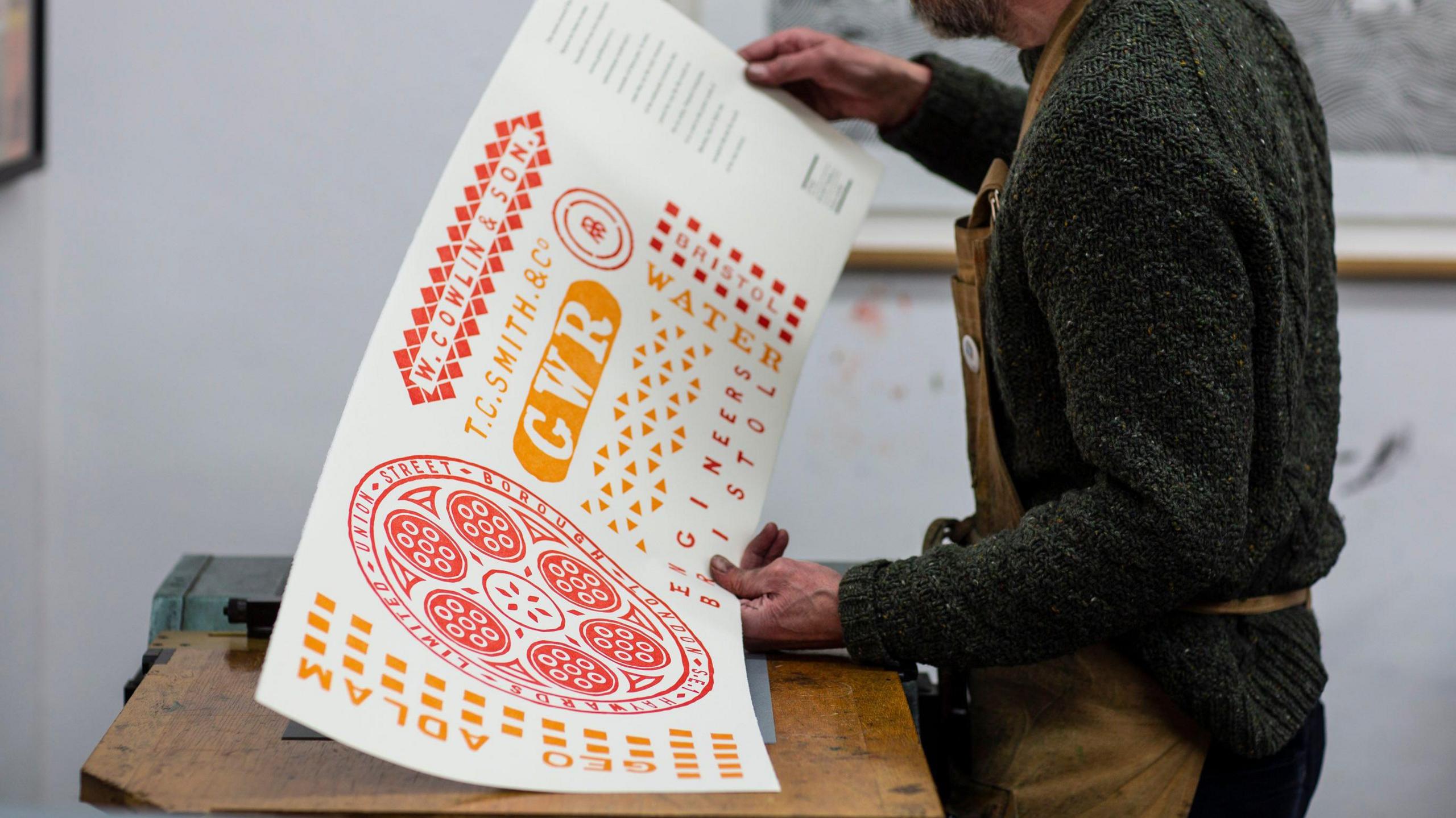 A man wearing a green knitted jumper and brown apron, holding up a commissioned piece of letter press printing. It is a large rectangular sign with red and orange GWR branding on it.