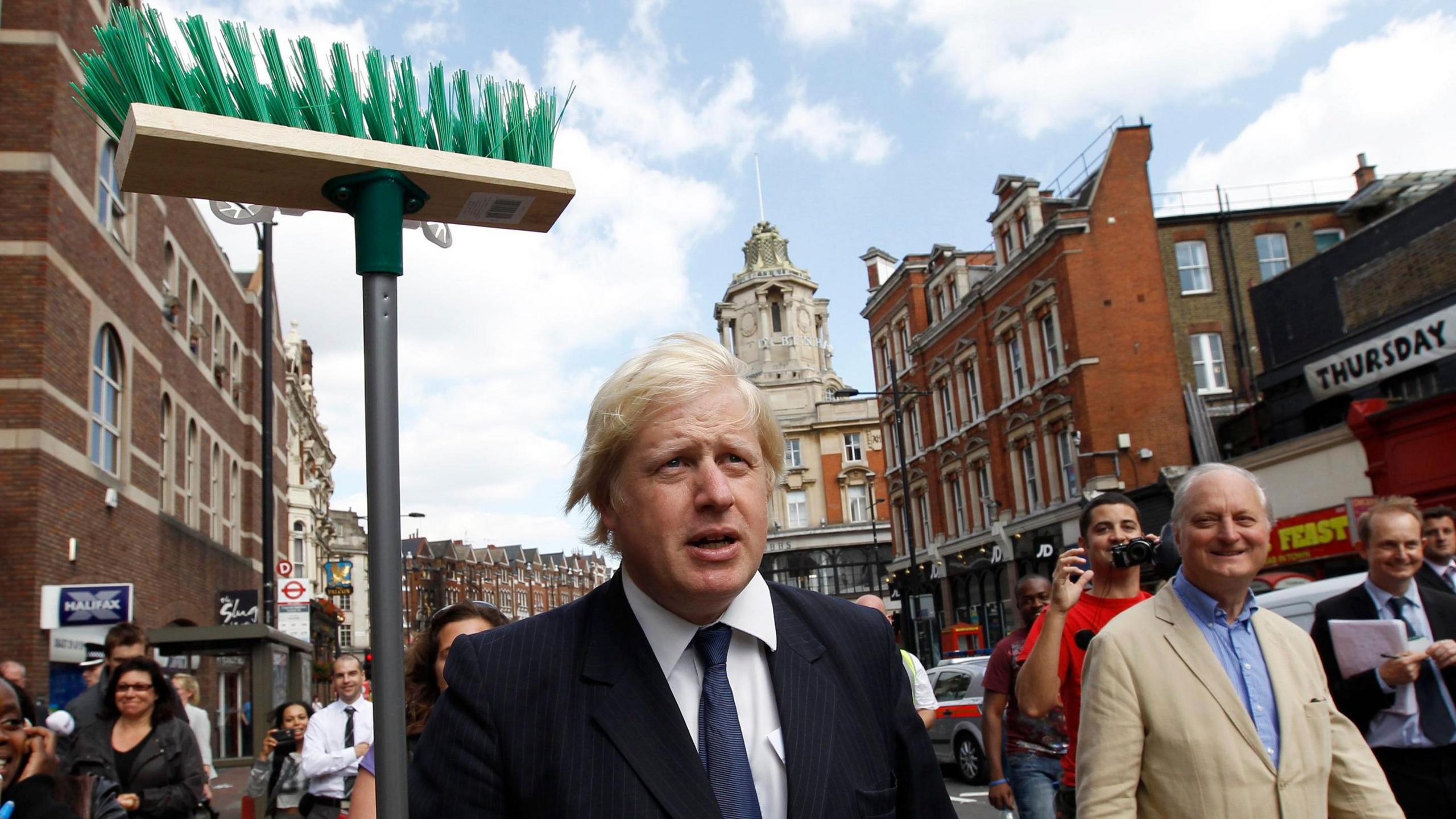 Boris Johnson, wearing a dark suit, white shirt and blue tie, stands on a London street with people behind him. He is holding aloft a broom with plastic green bristles. 