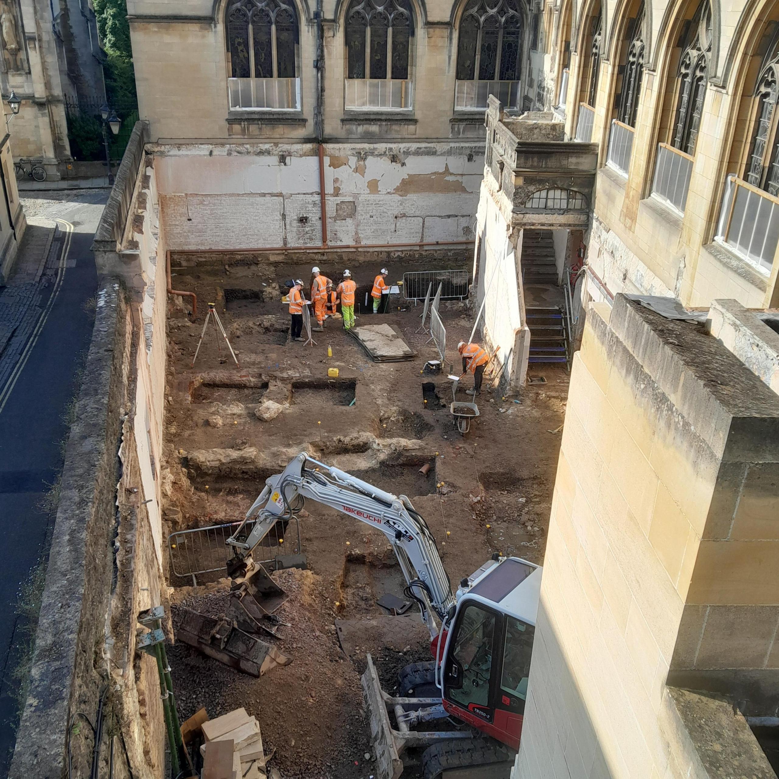 Excavated ground at the college, with squares carved into the soil. People in hard hats and an excavator vehicle can be seen.