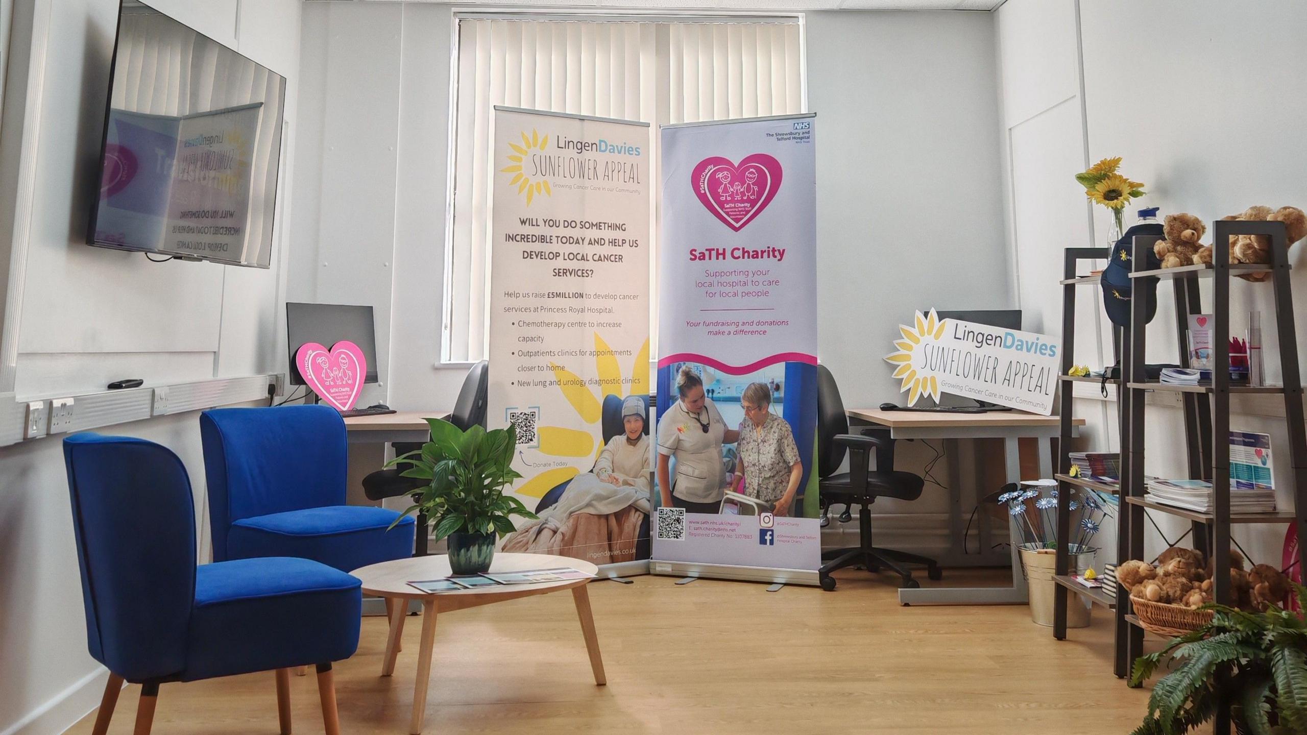 A room with two blue chairs and a small wooden table with a potted plant. Behind it on a wall is a television screen. At the back of the room is a computer on a desk. There are two large advertising stands, one reads "Lingen Davies Sunflower appeal" and the other reads "SaTH charity". on the right hand side of the room are two ladder-style shelves.