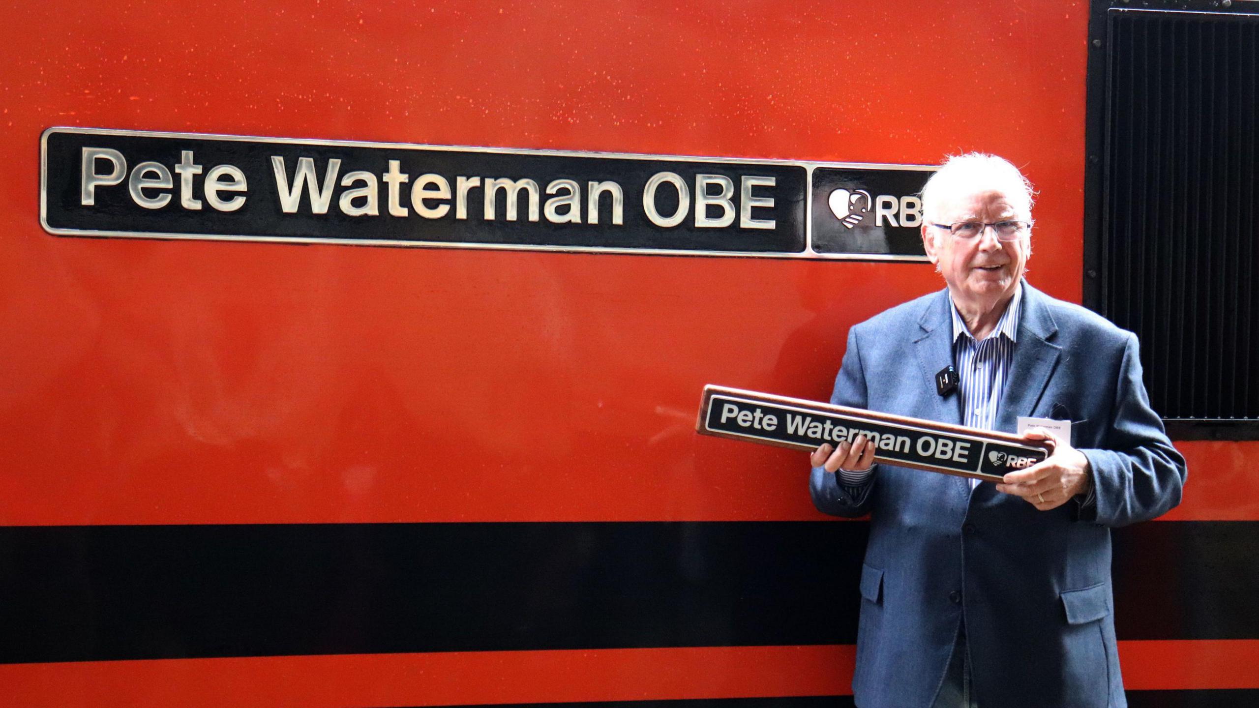 Pete Waterman standing next to a locomotive bearing his name. It is red and black and the sign on it says Pete Waterman OBE. 