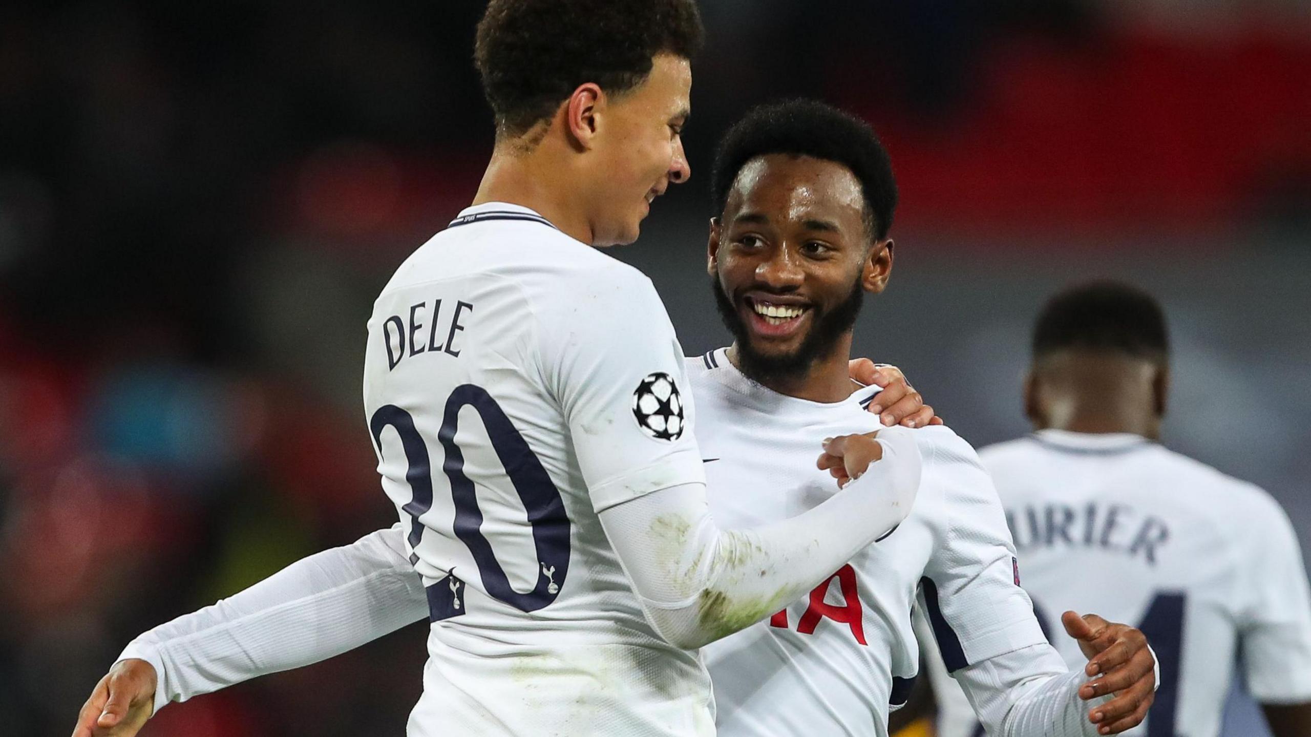 Dele Alli, with most of his back turned to the camera, hugs smiling Tottenham team-mate Georges-Kevin Nkoudou as fellow player Serge Aurier is shown in the background