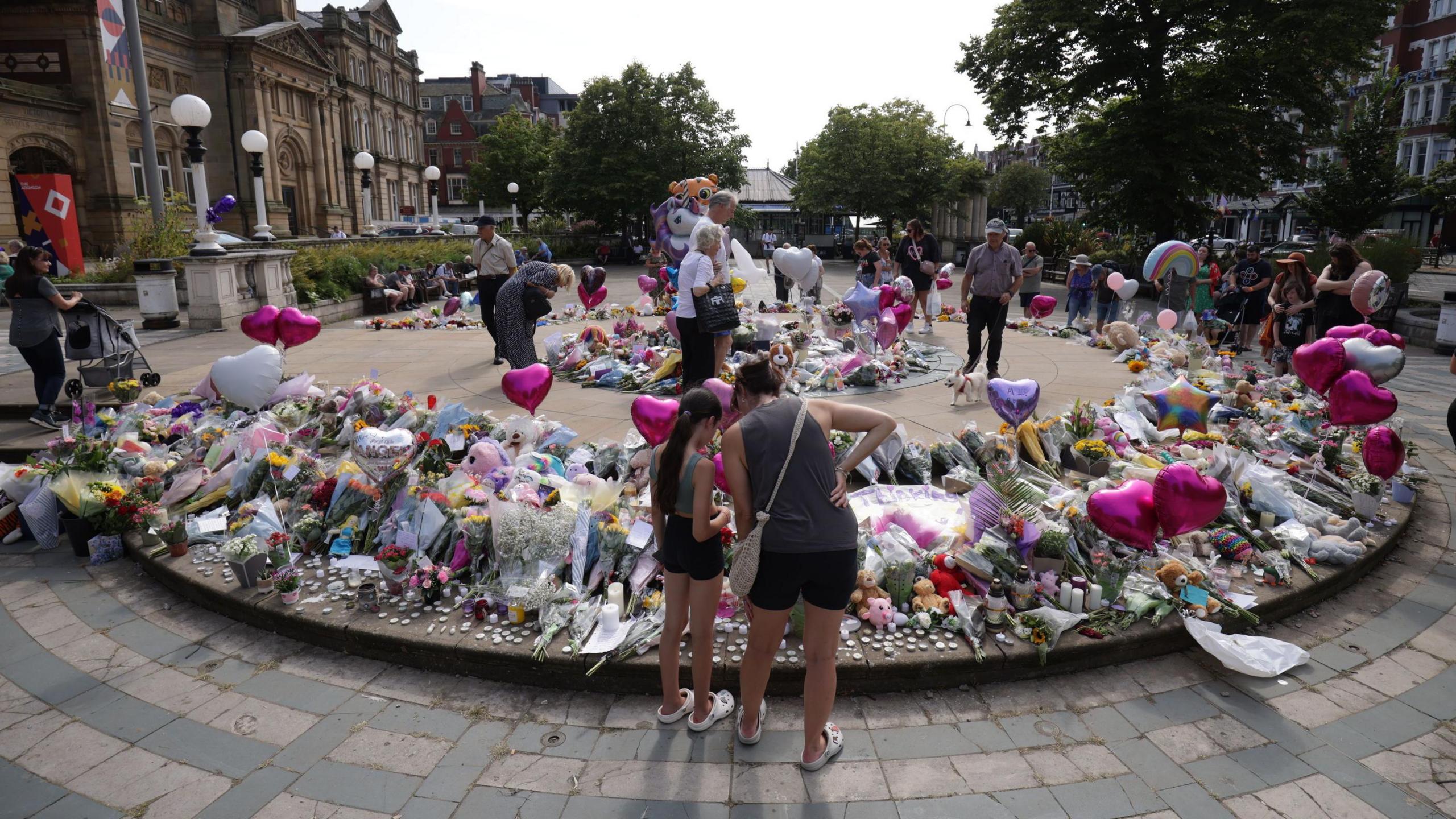 People stand and look at flowers, balloons, cuddly toys and other tributes left outside the Atkinson art centre in Southport