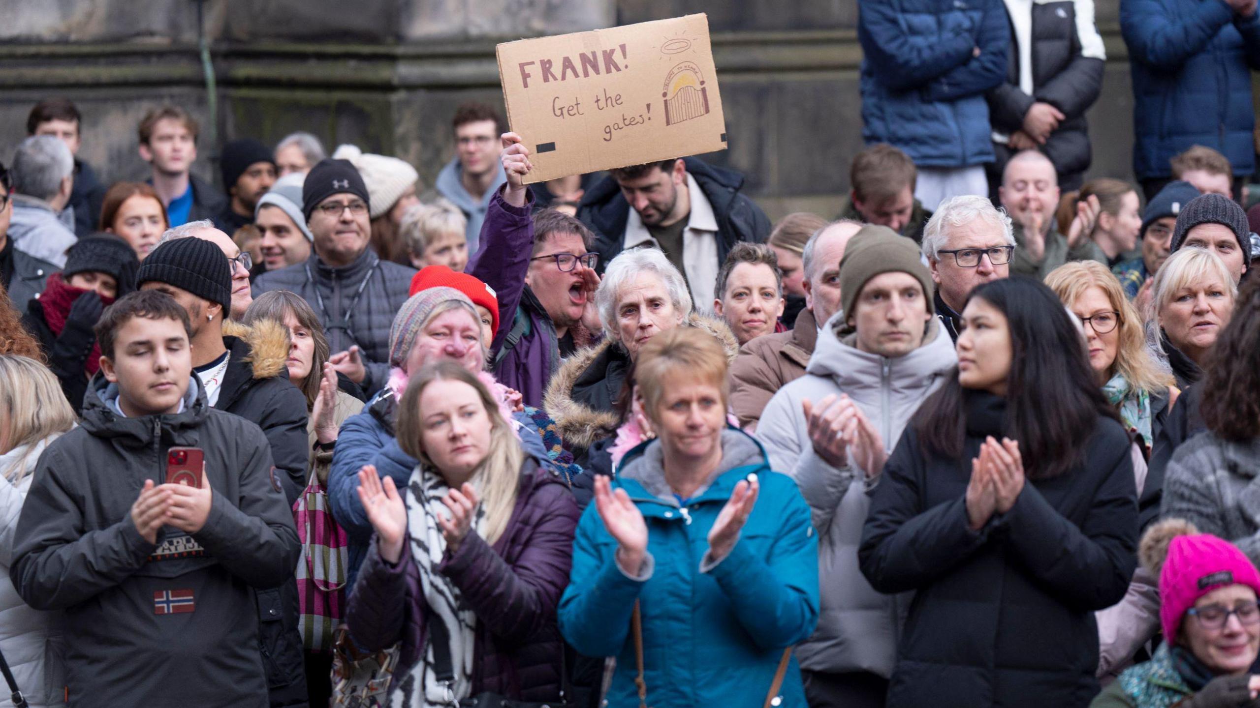 Dozens of mourners applauding as Janey Godley's hearse passes by. One of them is holding up a sign on cardboard saying 'Frank! Get the gates' with a drawing of gates.