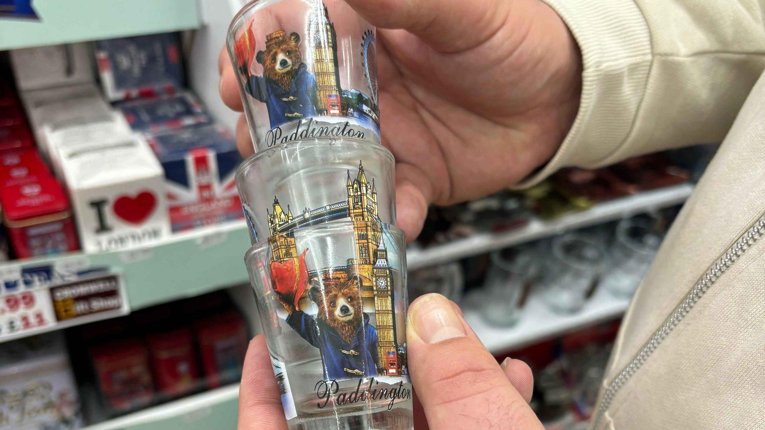 A man holds a stack of three glass shot glasses which have an artistic drawing of paddington bear waving his red hat, as well as Big Ben and Tower Bridge on them. Shelves of shop merchandise can be seen in the background